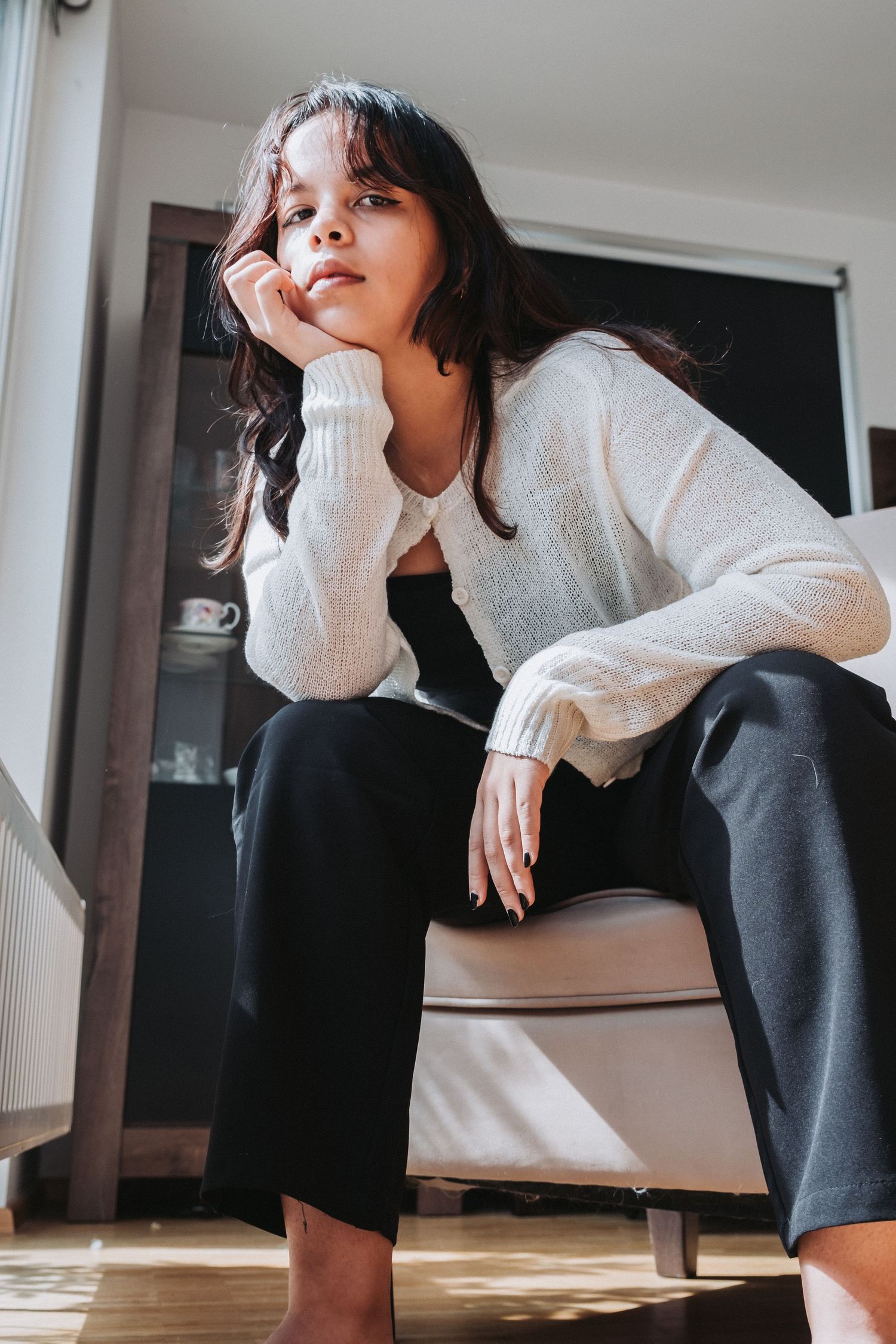 Woman in white sweater sitting thoughtfully on chair in sunlit room.
