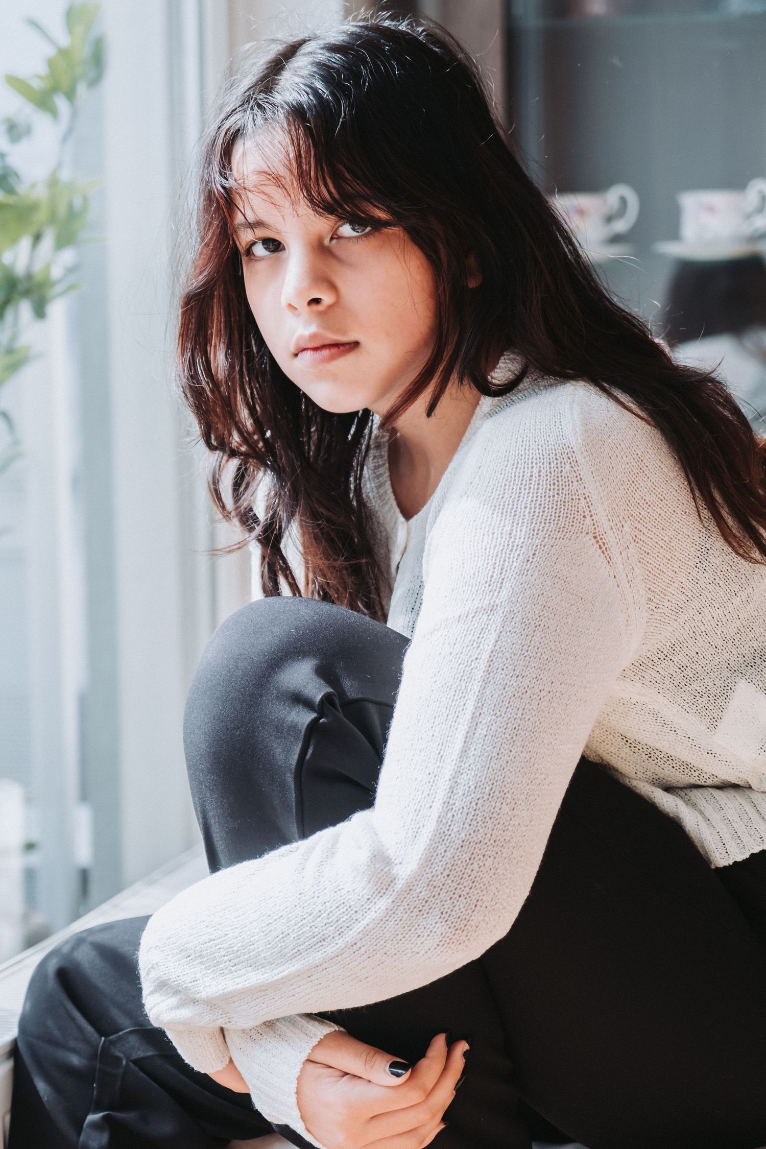 Person in white sweater sits by window in casual indoor portrait.