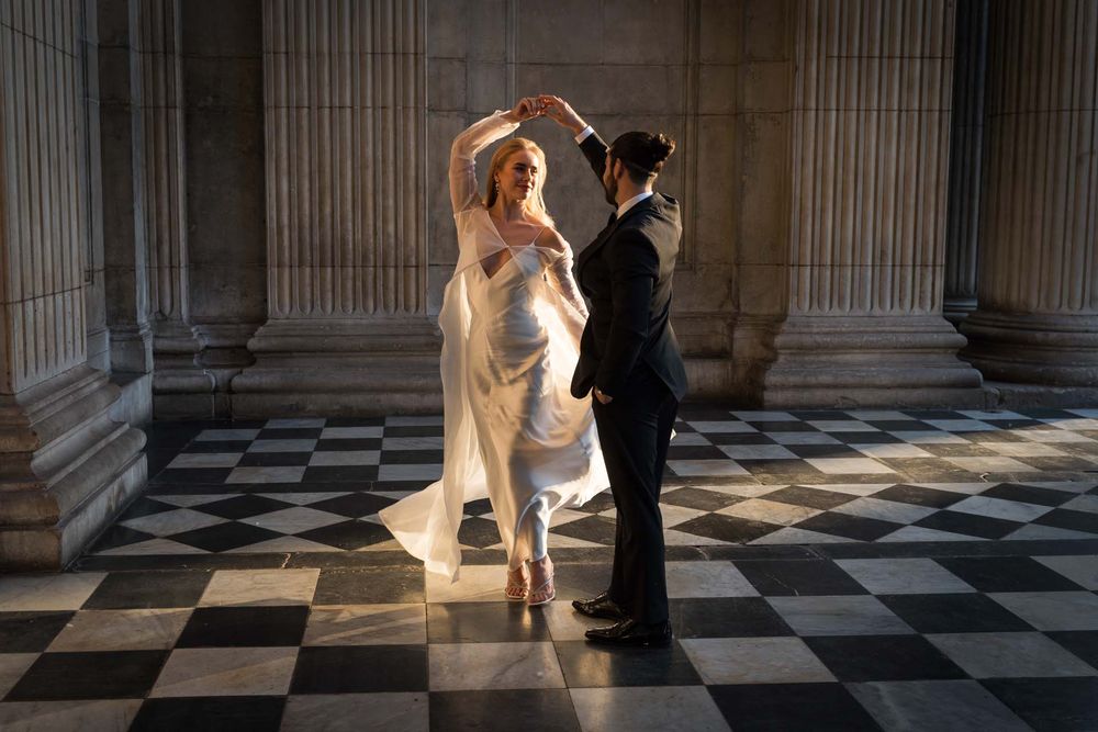 A couple dances elegantly on a black and white checkered floor surrounded by classic architecture.
