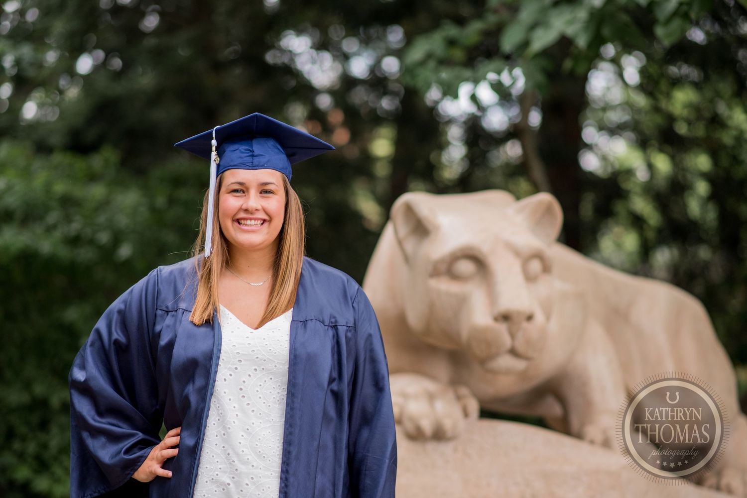 Smiling graduate in blue regalia stands beside iconic Penn State lion sculpture.