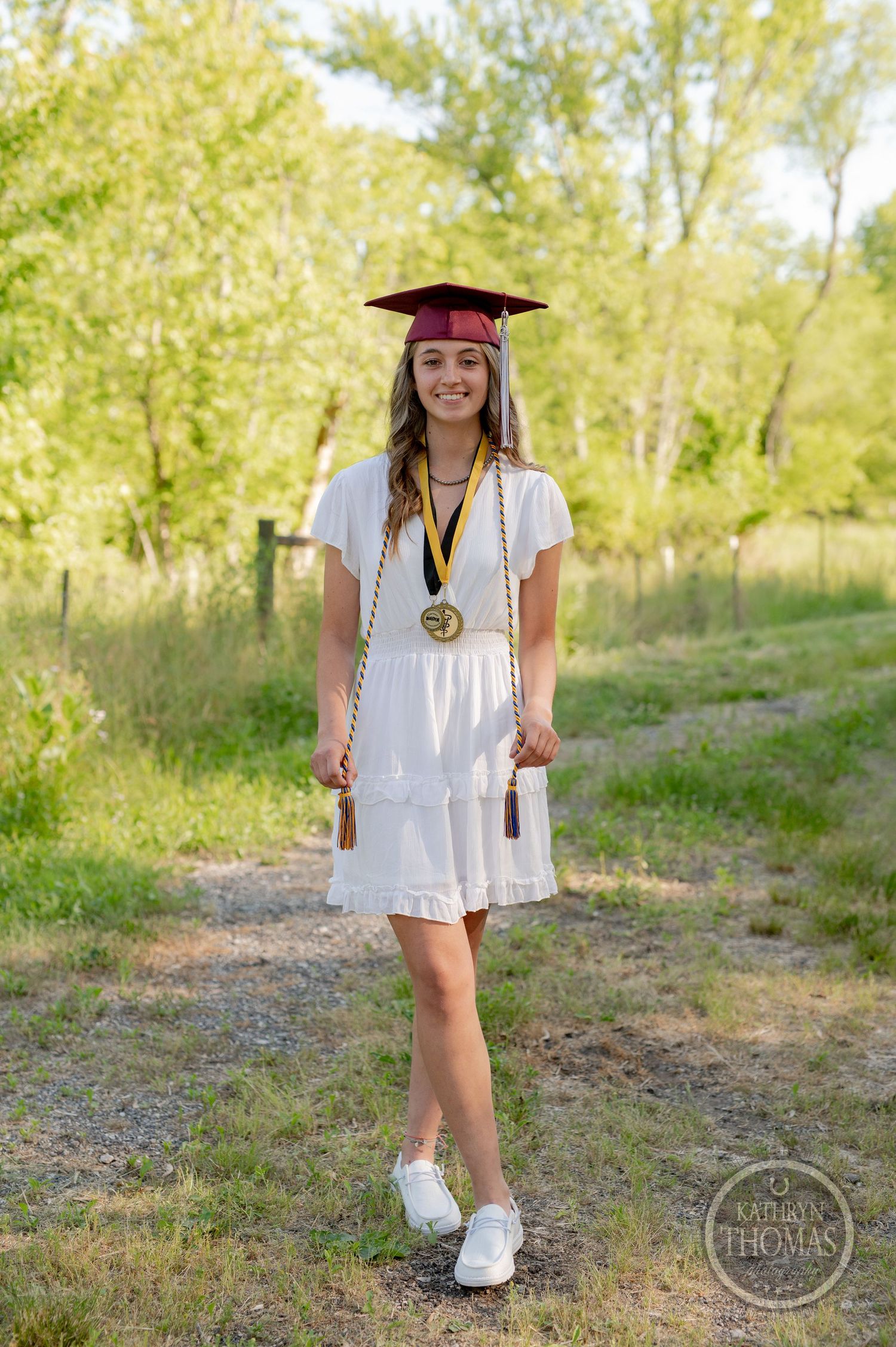 Graduate wearing white dress and maroon cap stands on wooded path with medal and graduation regalia.