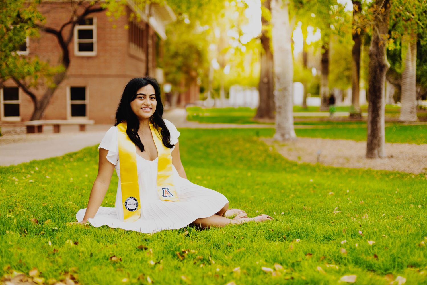 Graduate in white dress poses on grass lawn near campus buildings.