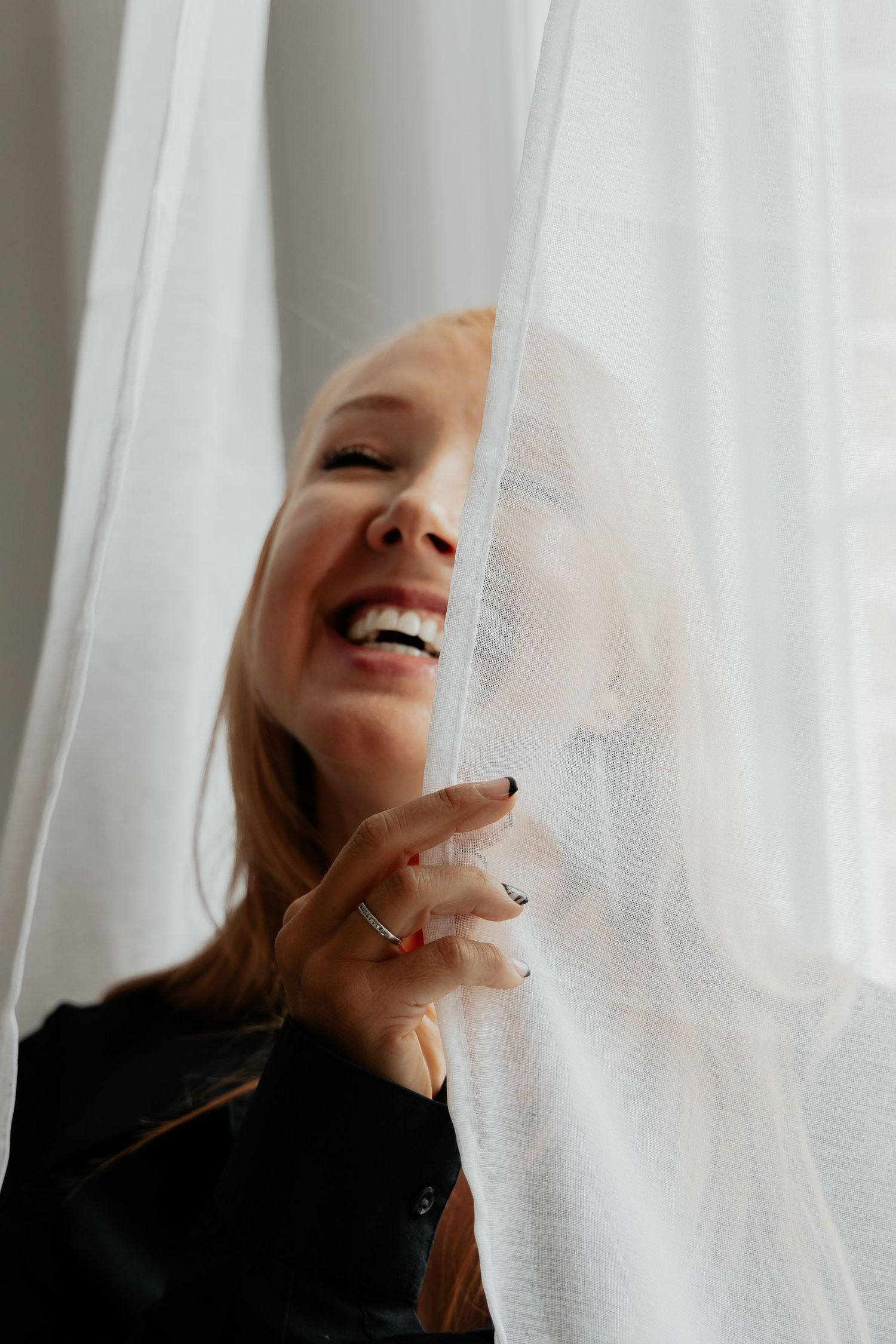 Joyful woman laughing behind sheer white curtains with natural window light.