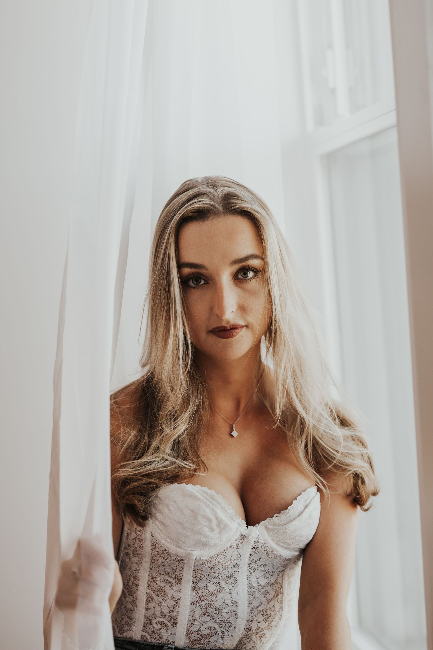 Blonde woman in white lace corset standing by sheer curtained window in soft natural light.