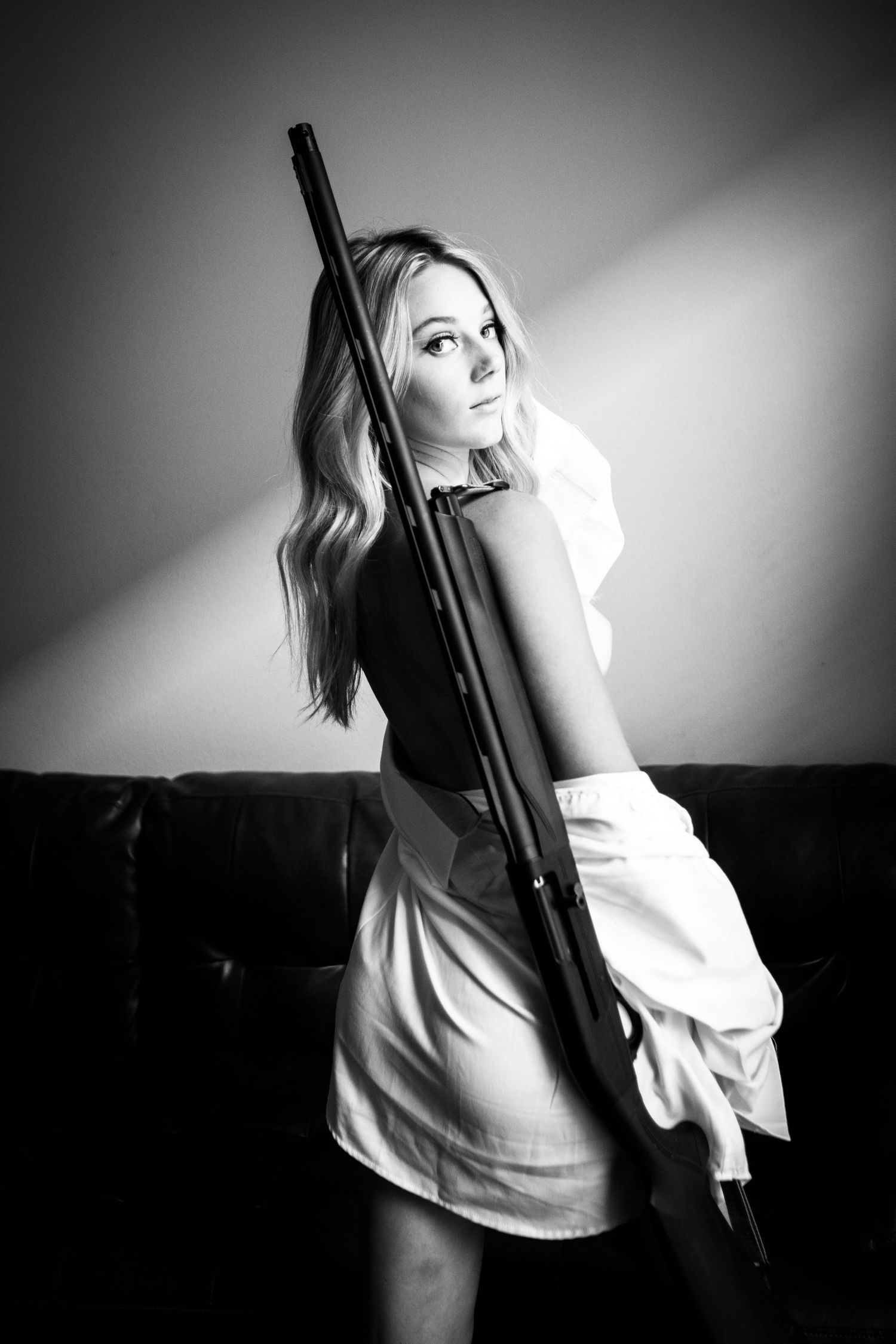 Black and white portrait of blonde woman holding a rifle against her back in a white shirt.
