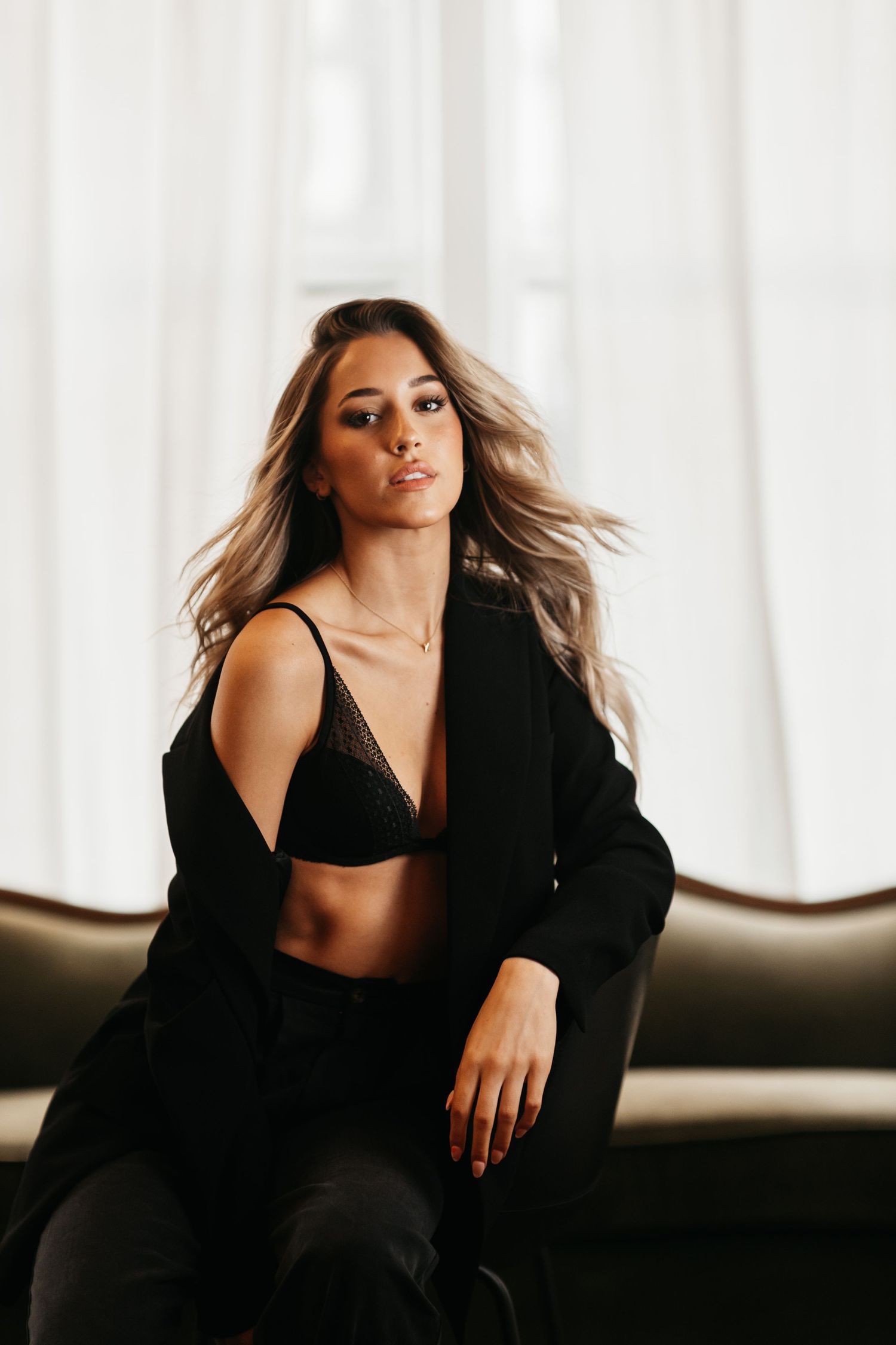 Young woman in black bra and oversized blazer sitting on dark sofa near white curtains.