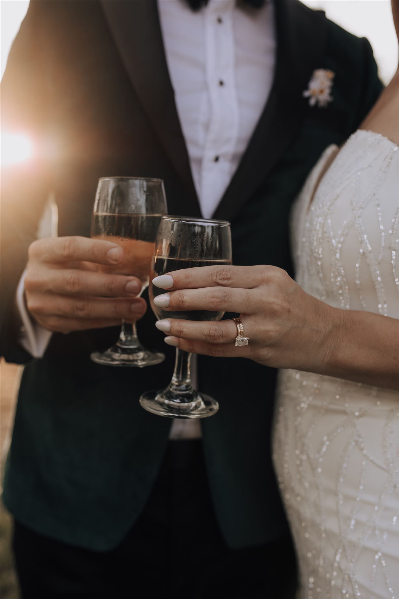 A couple toasts with wine glasses, showing off wedding rings and formal attire.