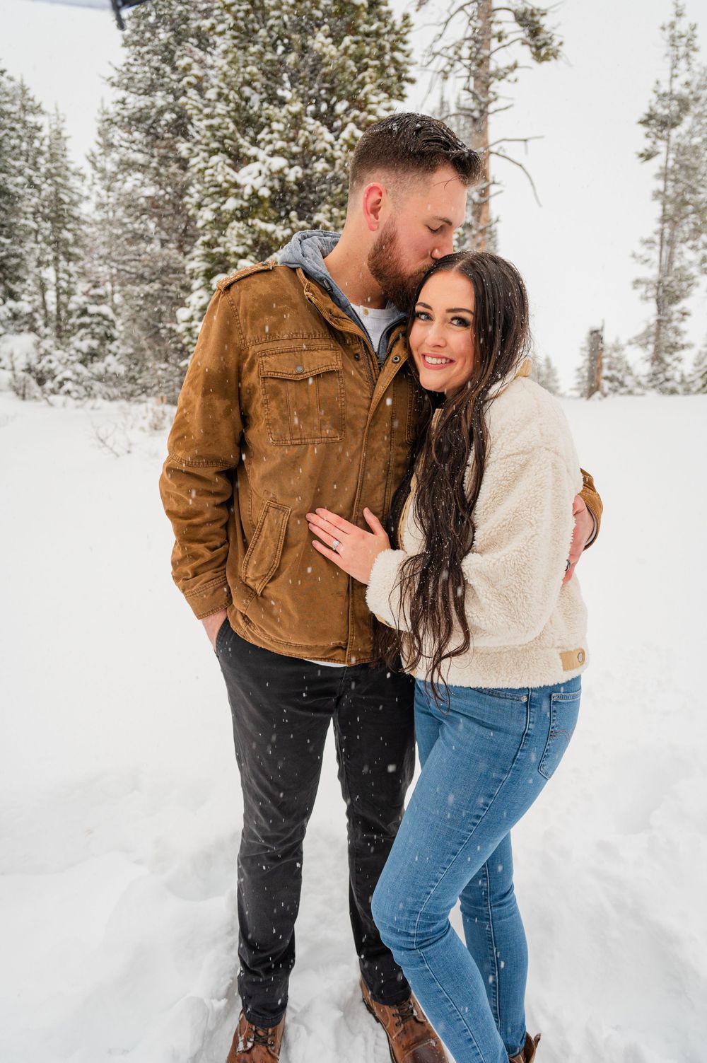 Couple poses together in a snowy forest setting wearing winter casual attire.