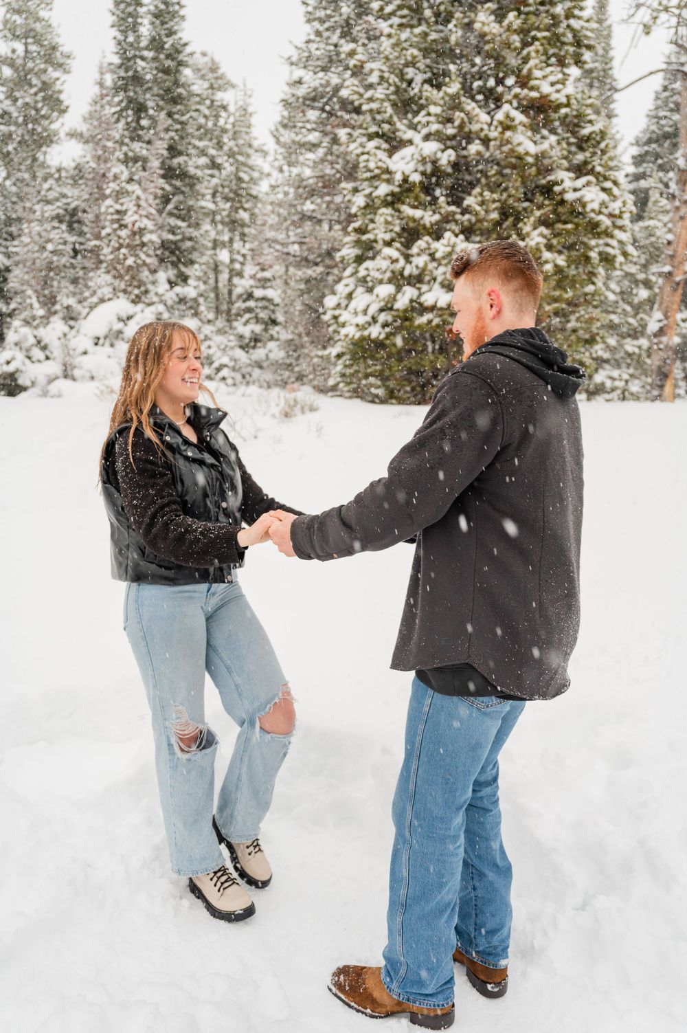 Couple holding hands and dancing in the snow with evergreen trees in the background.