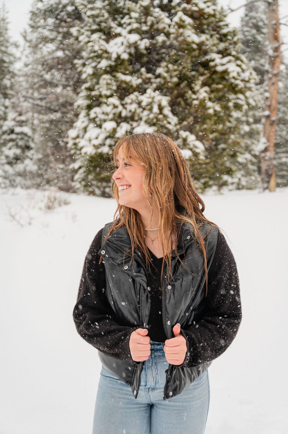 A person wearing a black jacket and jeans smiles outdoors in a snowy winter forest setting.