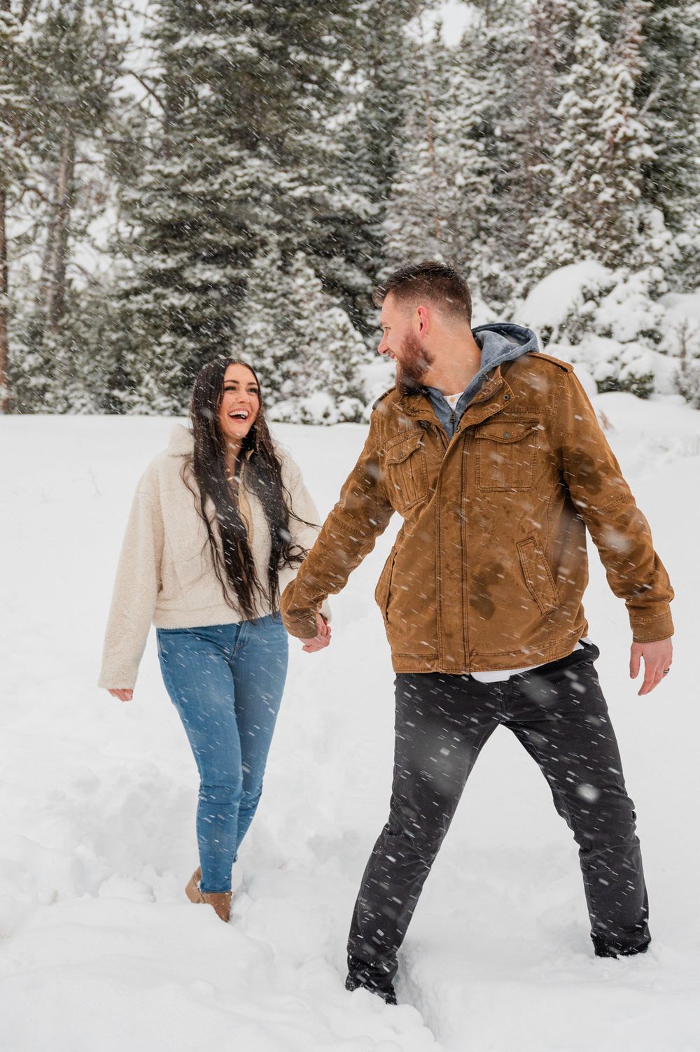 A couple walking hand in hand through a snowy forest, wearing winter jackets and jeans.