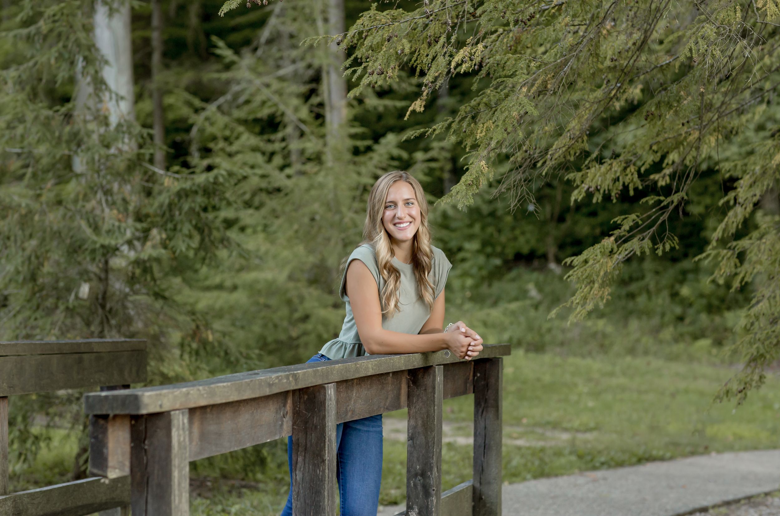 Smiling blonde teen leaning on wooden bridge railing surrounded by tall green pine trees.