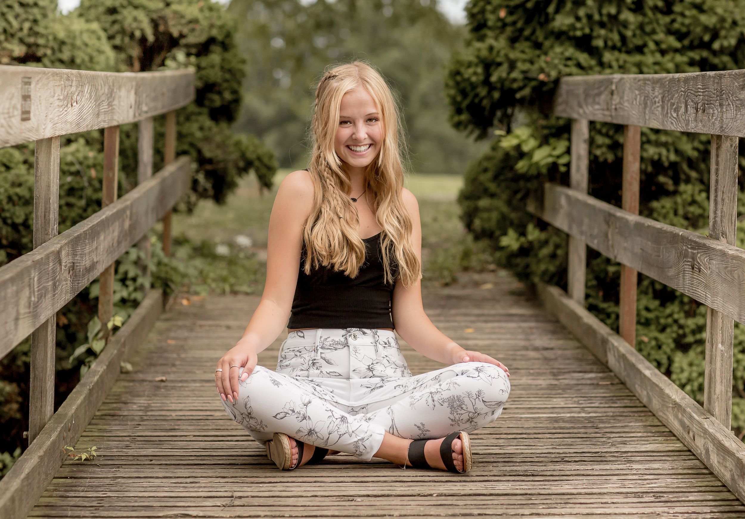 Smiling blonde teen in black top and white pants sitting cross-legged on a wooden bridge.