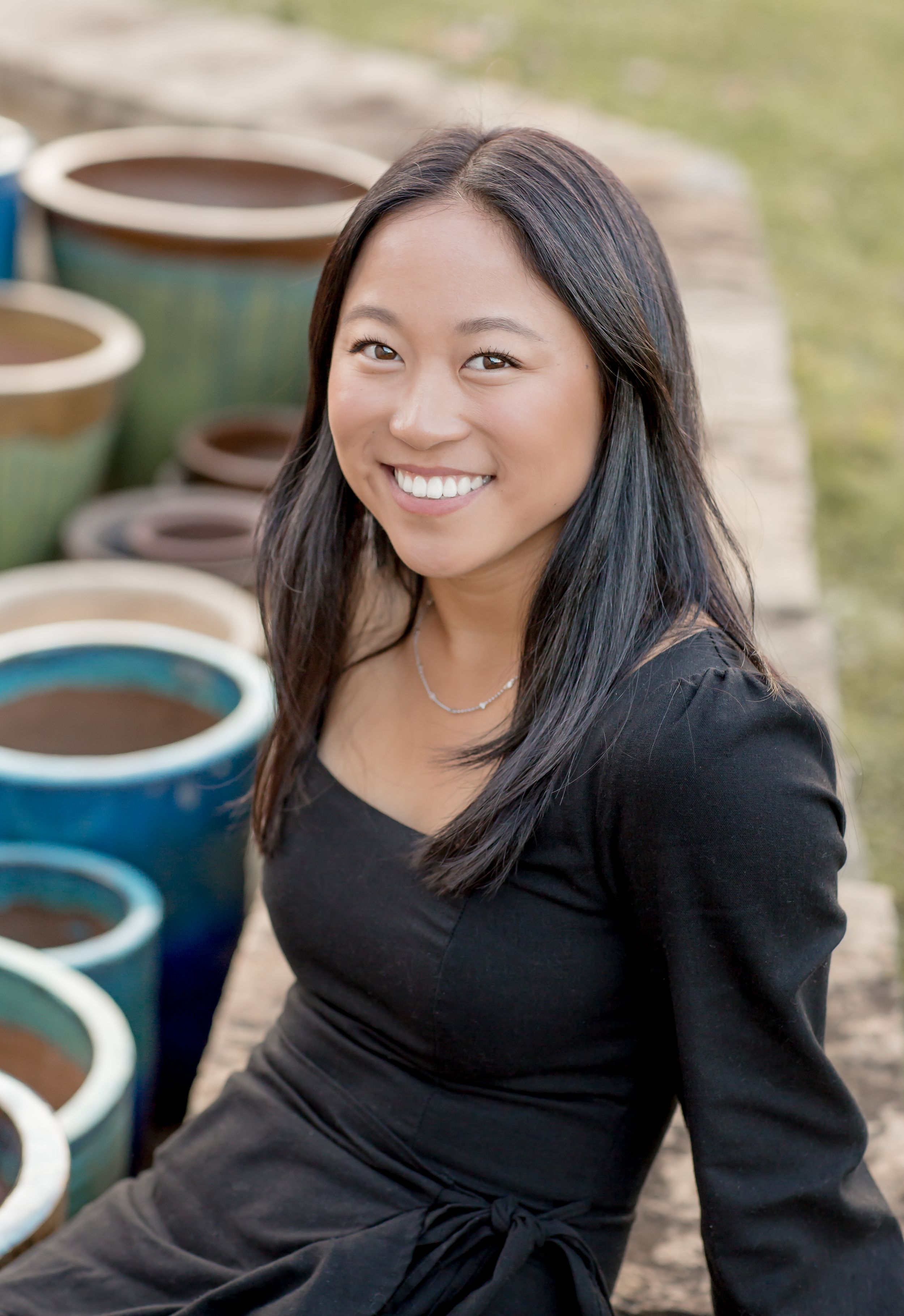 Smiling Asian woman in black wrap top sitting outdoors near colorful ceramic garden pots.