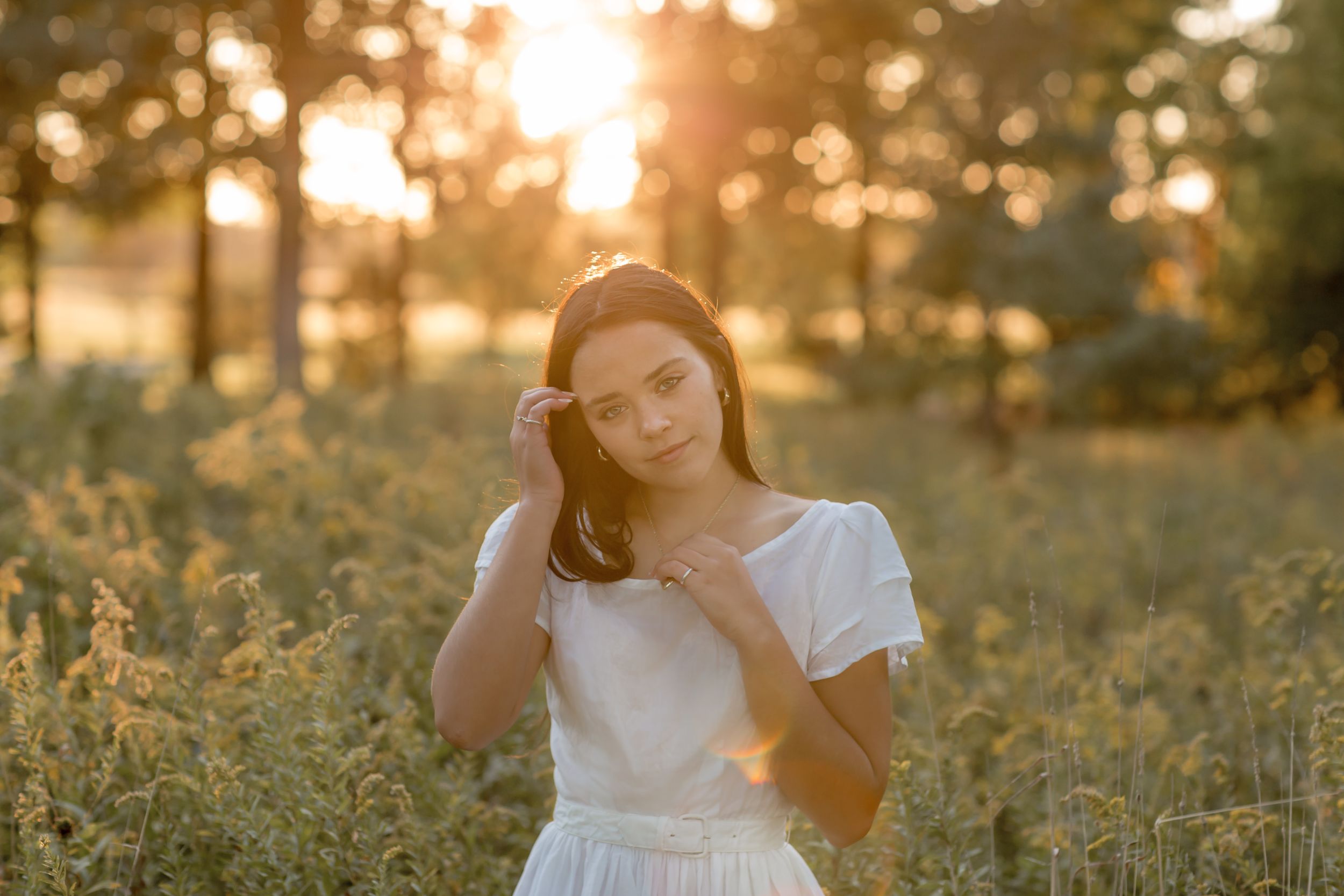 Young woman in white dress standing in a golden sunset field touching her hair.