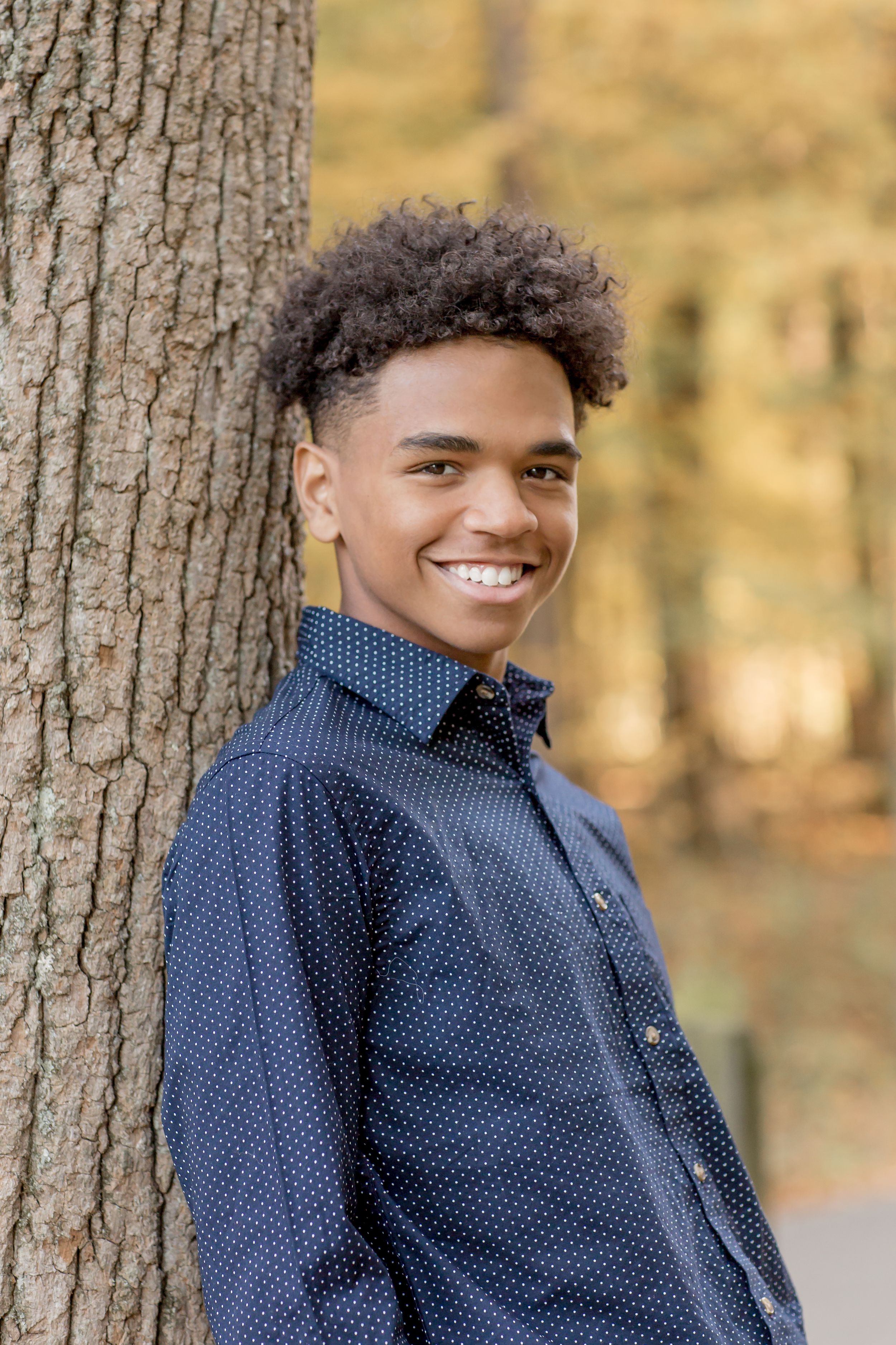 Smiling teenage boy in navy polka dot shirt leaning against a tree in autumn park.