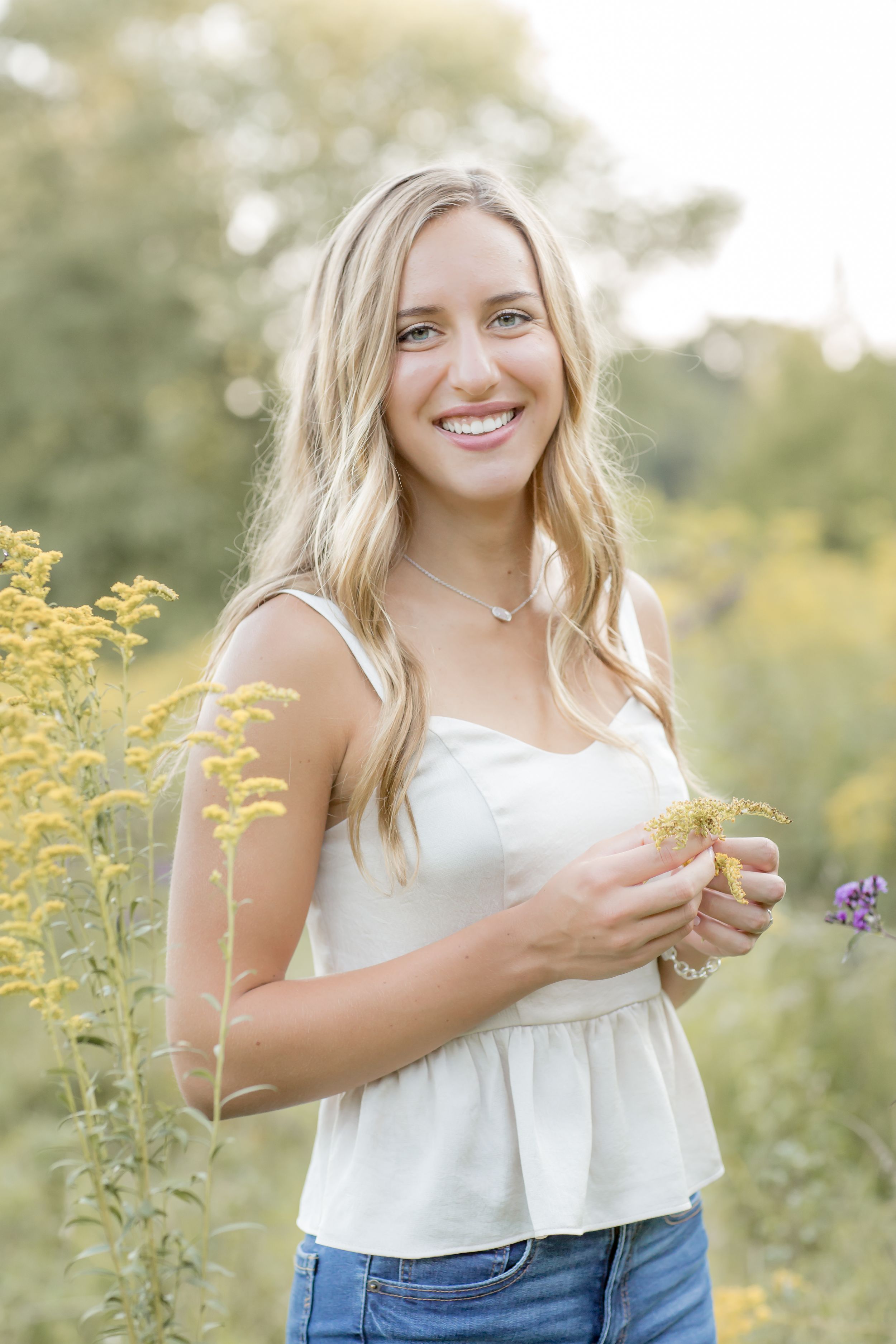 Smiling blonde teen in white top holding wildflowers in a sunny outdoor meadow senior portrait.