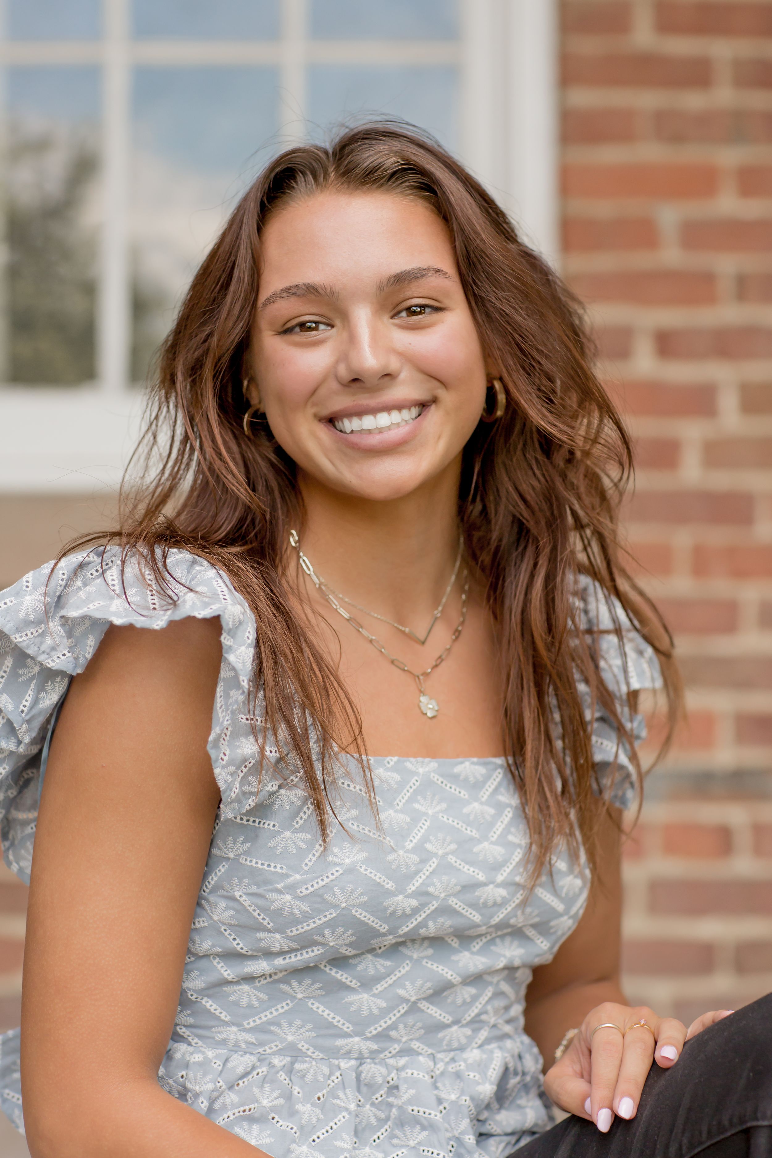Smiling young woman with long brown hair wearing a light blue ruffled top and layered necklaces outdoors.