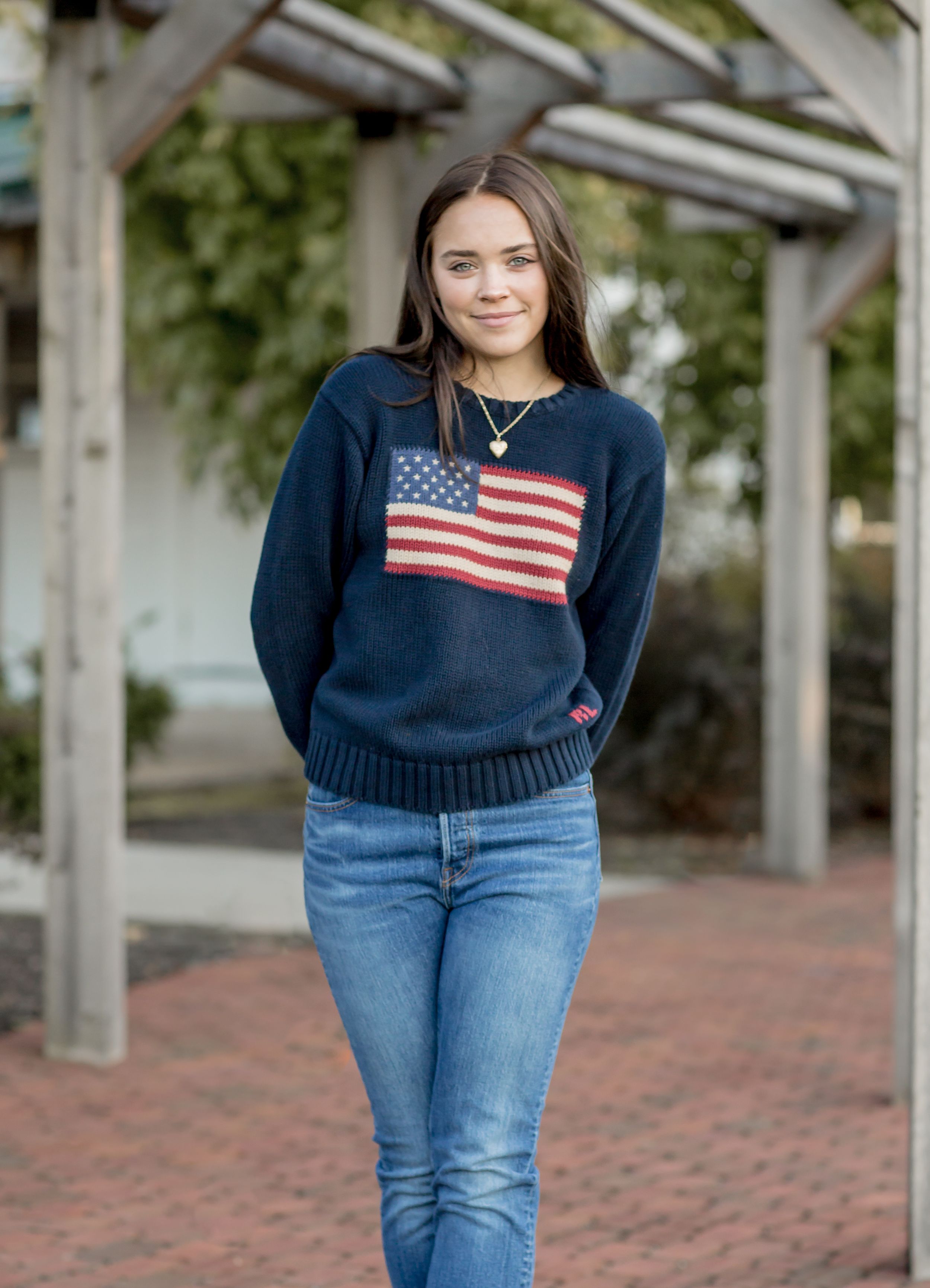 Young woman wearing a navy American flag sweater and jeans standing under a pergola.