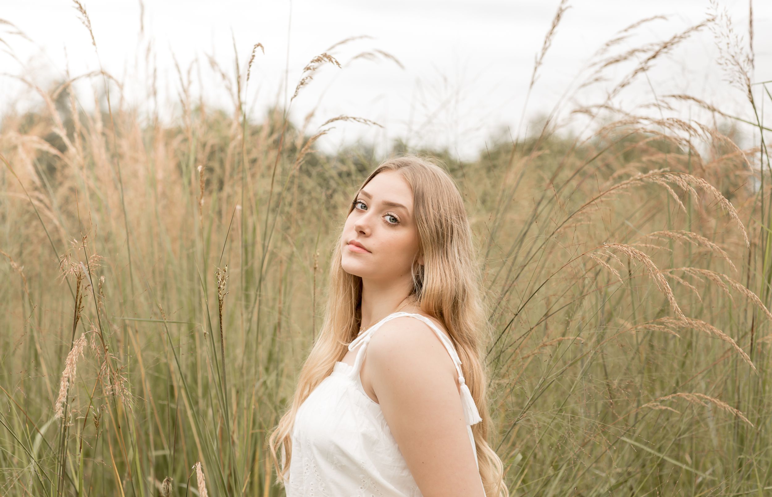 Young blonde woman in white dress standing among tall golden grass in outdoor senior photo.
