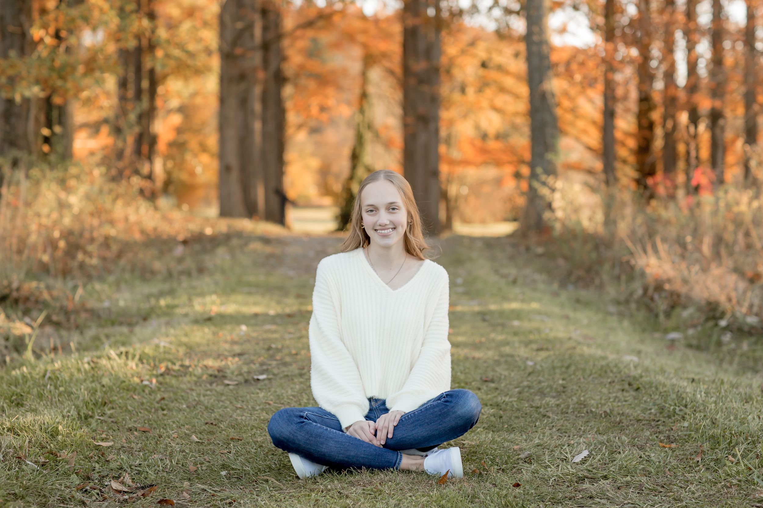 Smiling young blonde woman in a white sweater sitting cross-legged on a path in autumn forest.