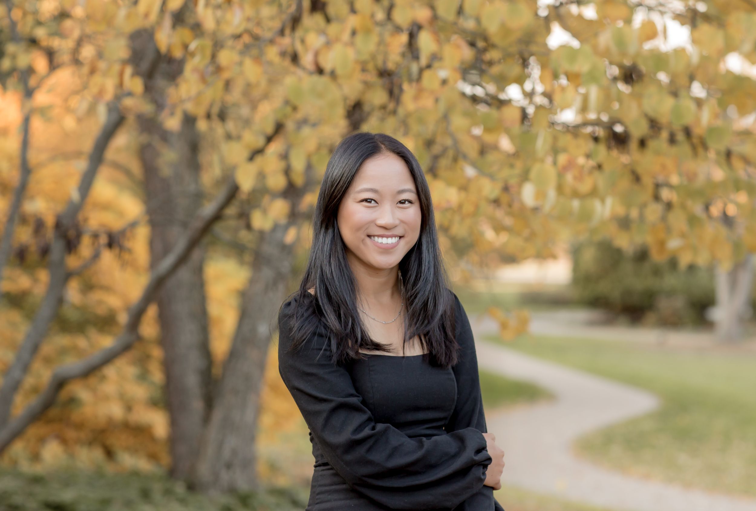 Smiling young Asian woman in black outfit standing outdoors among autumn foliage trees.