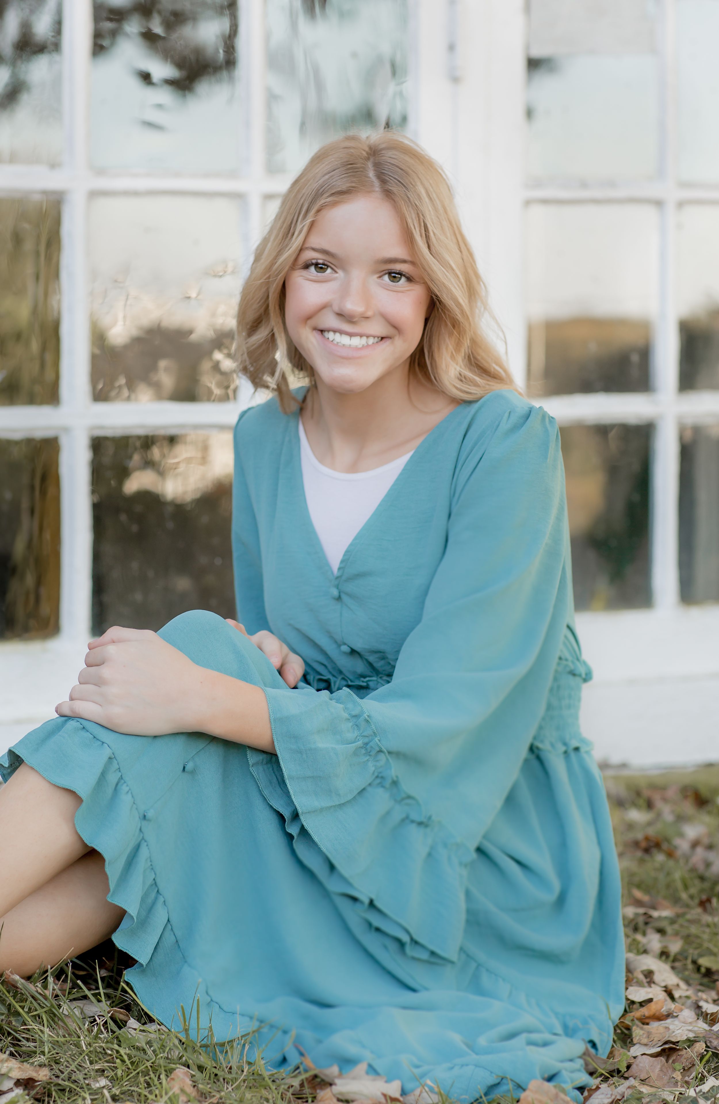 Smiling blonde teenage girl in teal ruffled dress sitting on grass near a window.