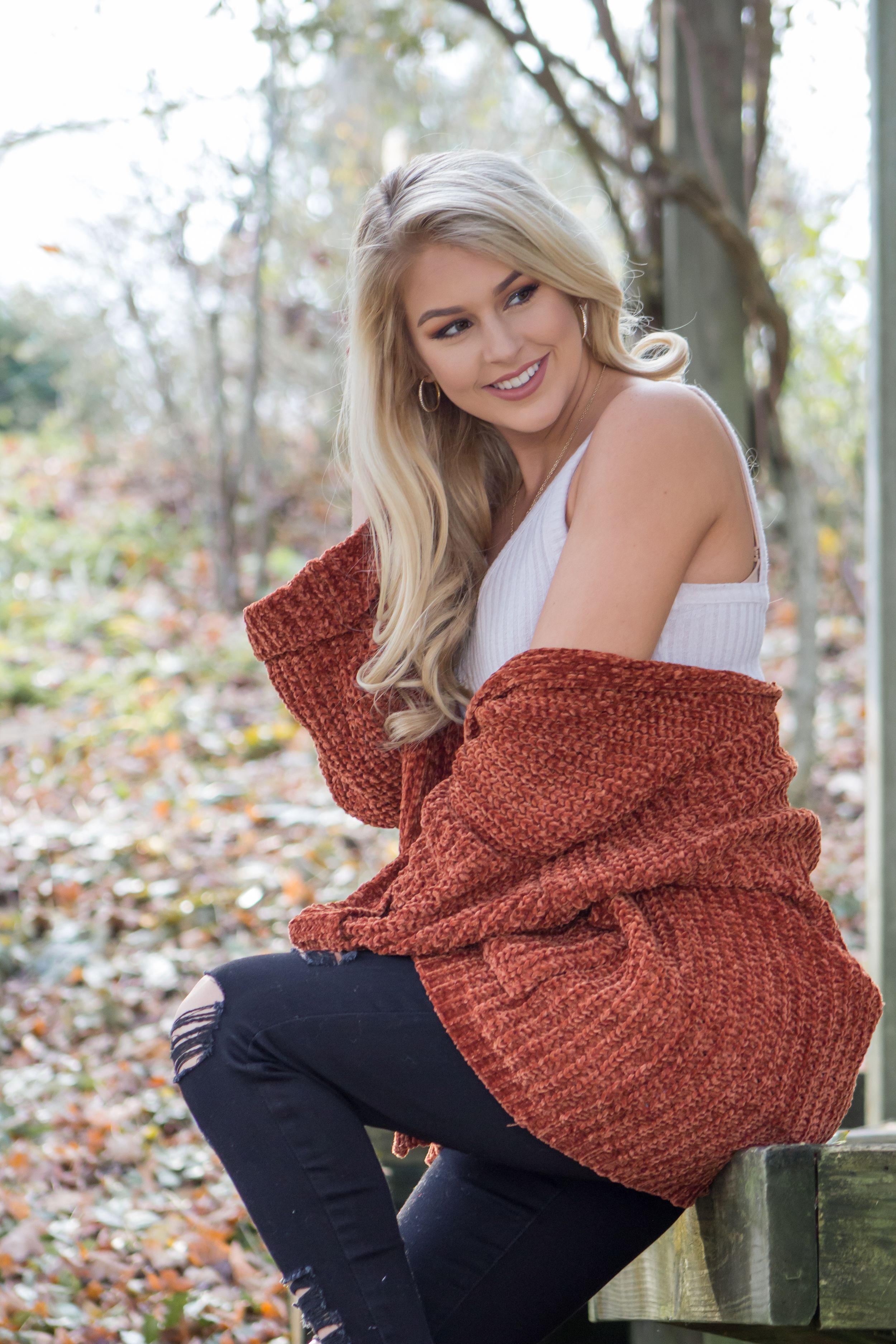 Young woman in rust orange knit cardigan sitting outdoors among autumn foliage.