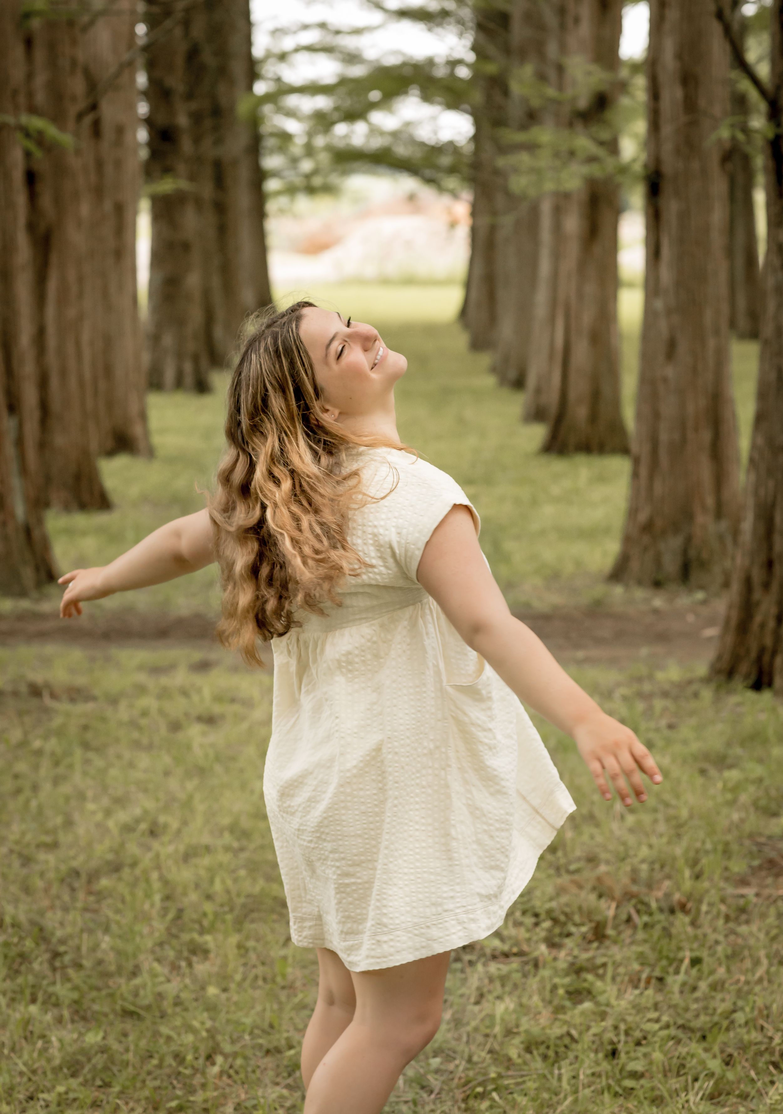 Young woman in a cream dress twirling joyfully between tall trees in a grassy outdoor setting.