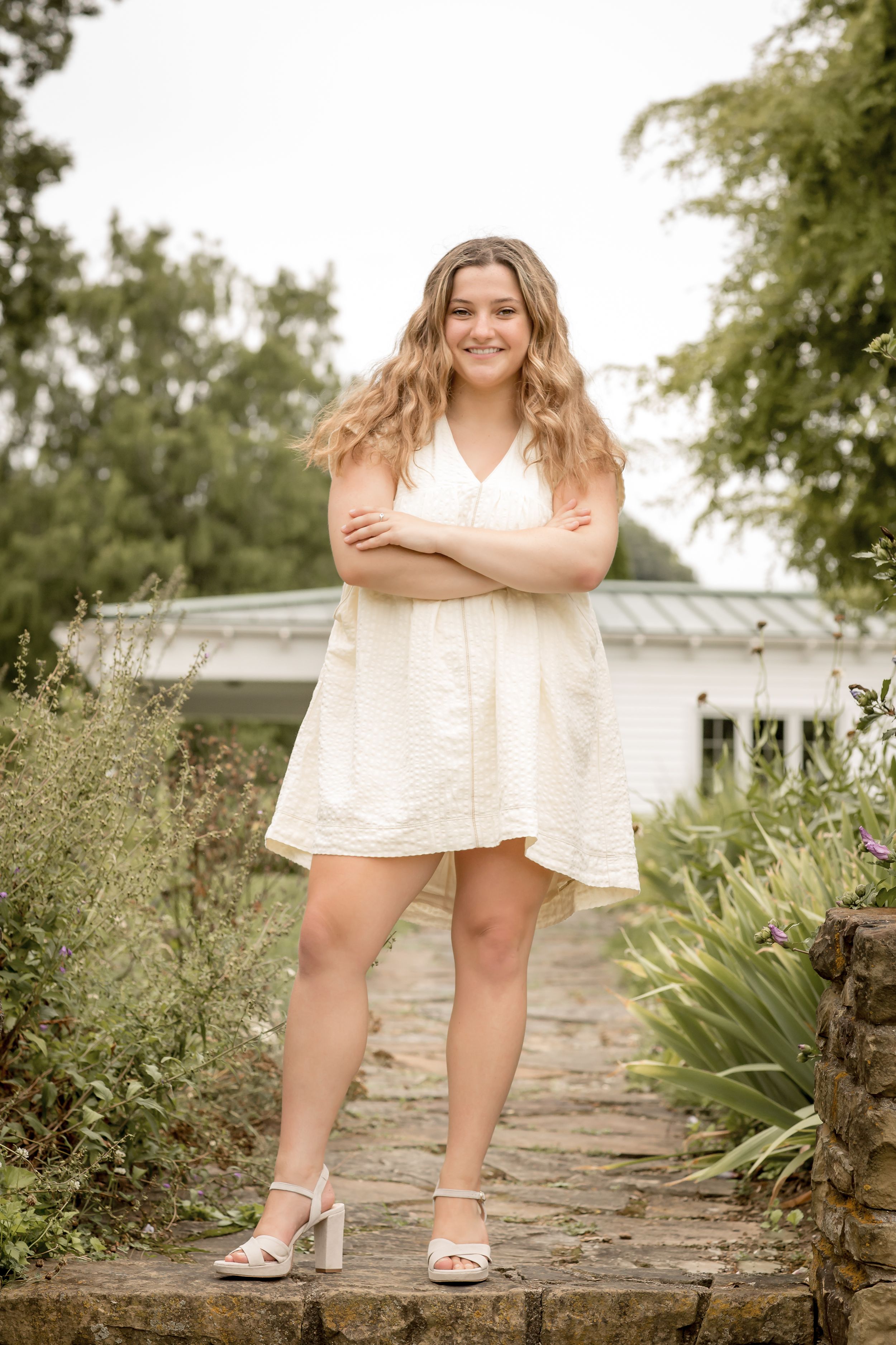 Smiling young woman in white mini dress with arms crossed standing on stone path in garden.