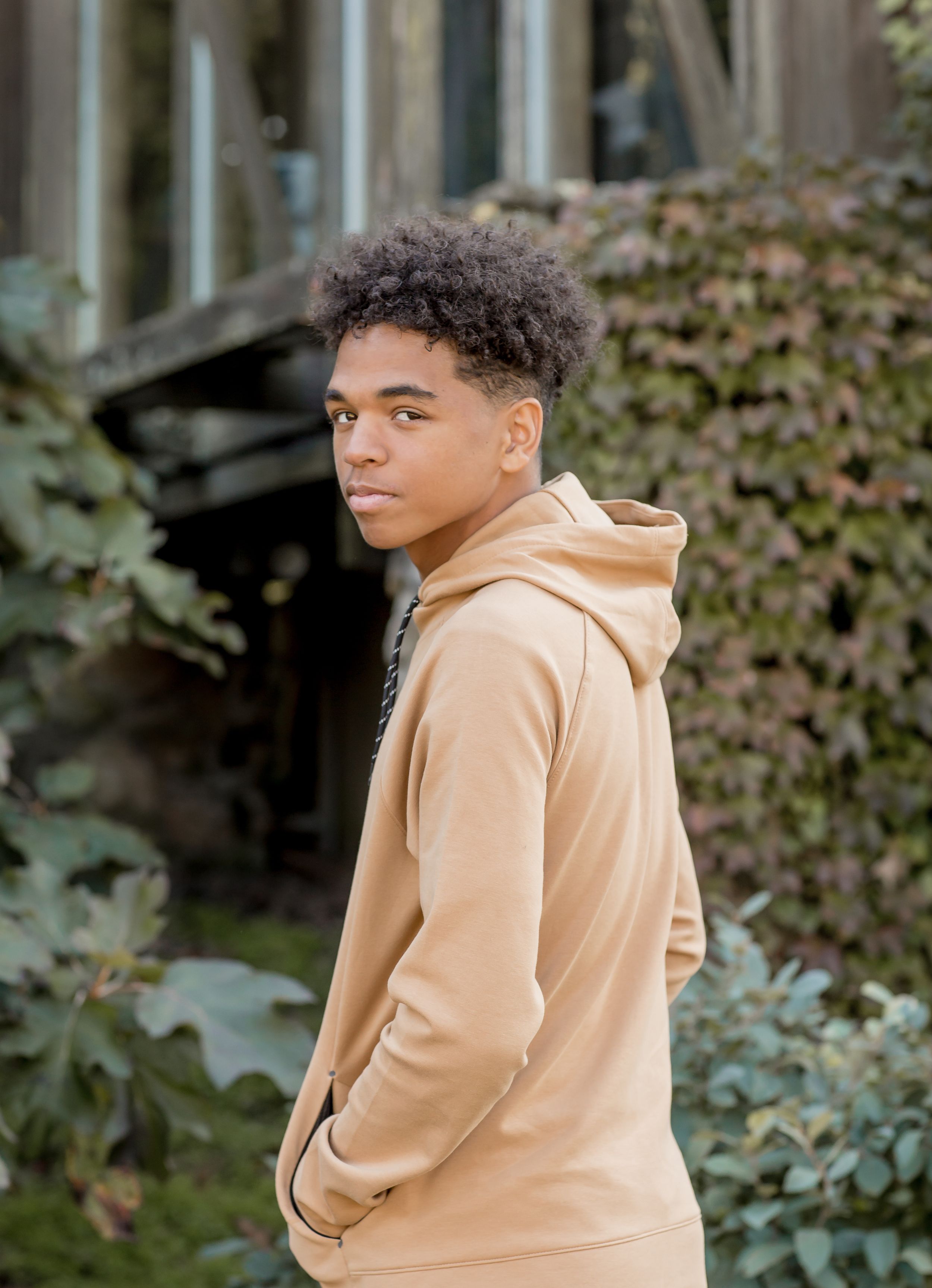 Teenage boy in tan hoodie looking over his shoulder in a garden with ivy backdrop.