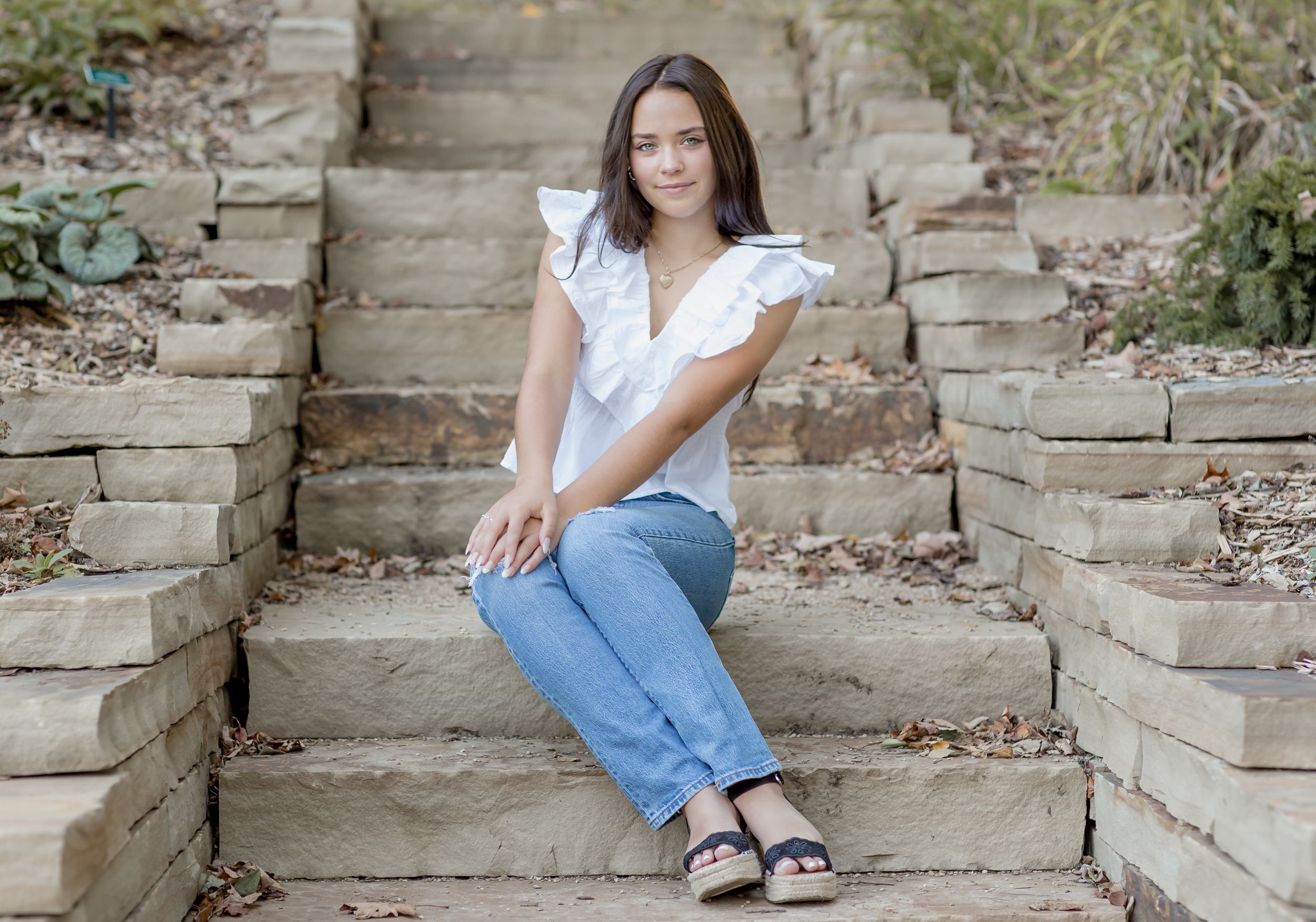 Young woman in white ruffled top and jeans sitting on stone steps in a garden setting.