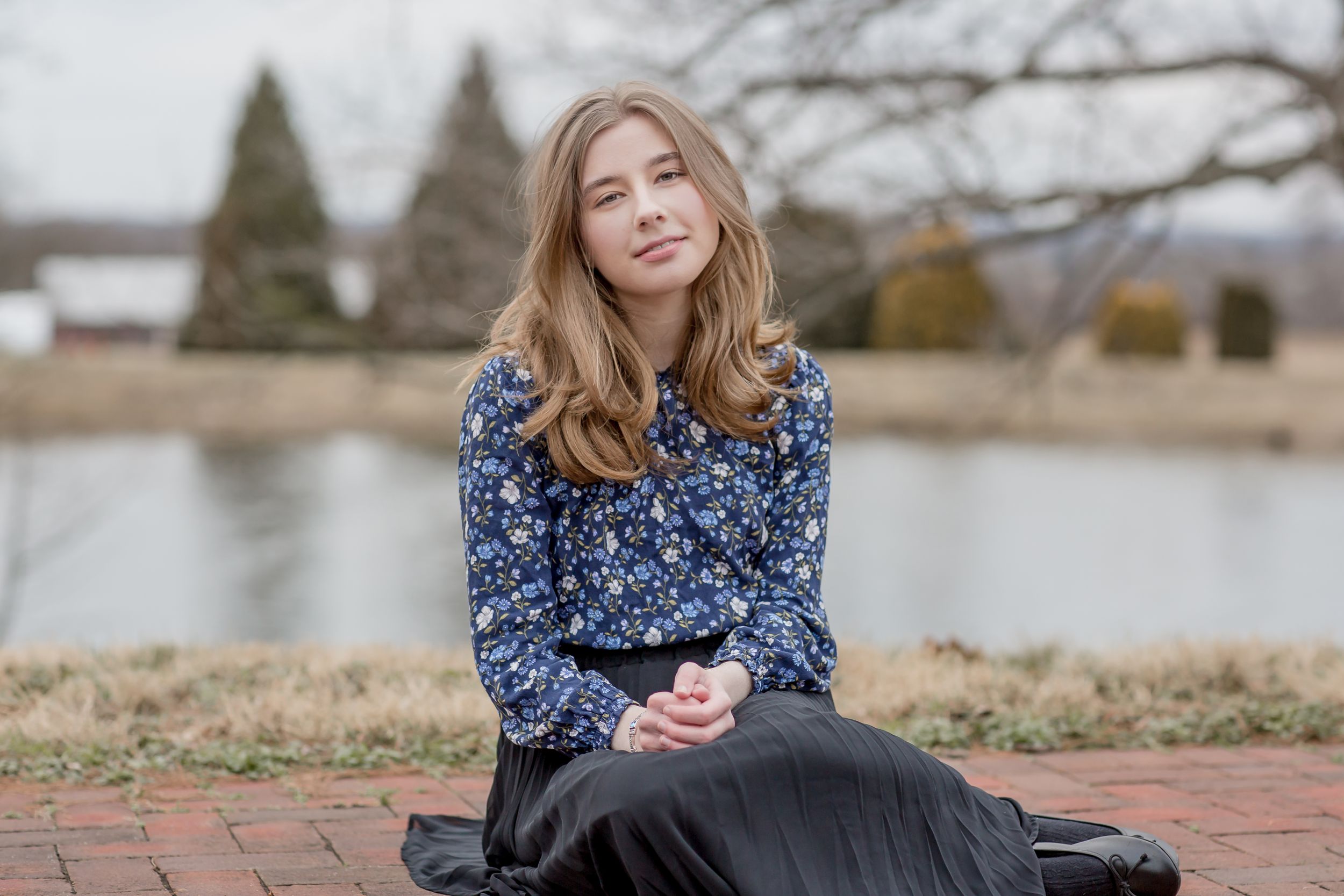 Young woman in a floral blouse and black skirt sitting on brick pavers near a serene pond.