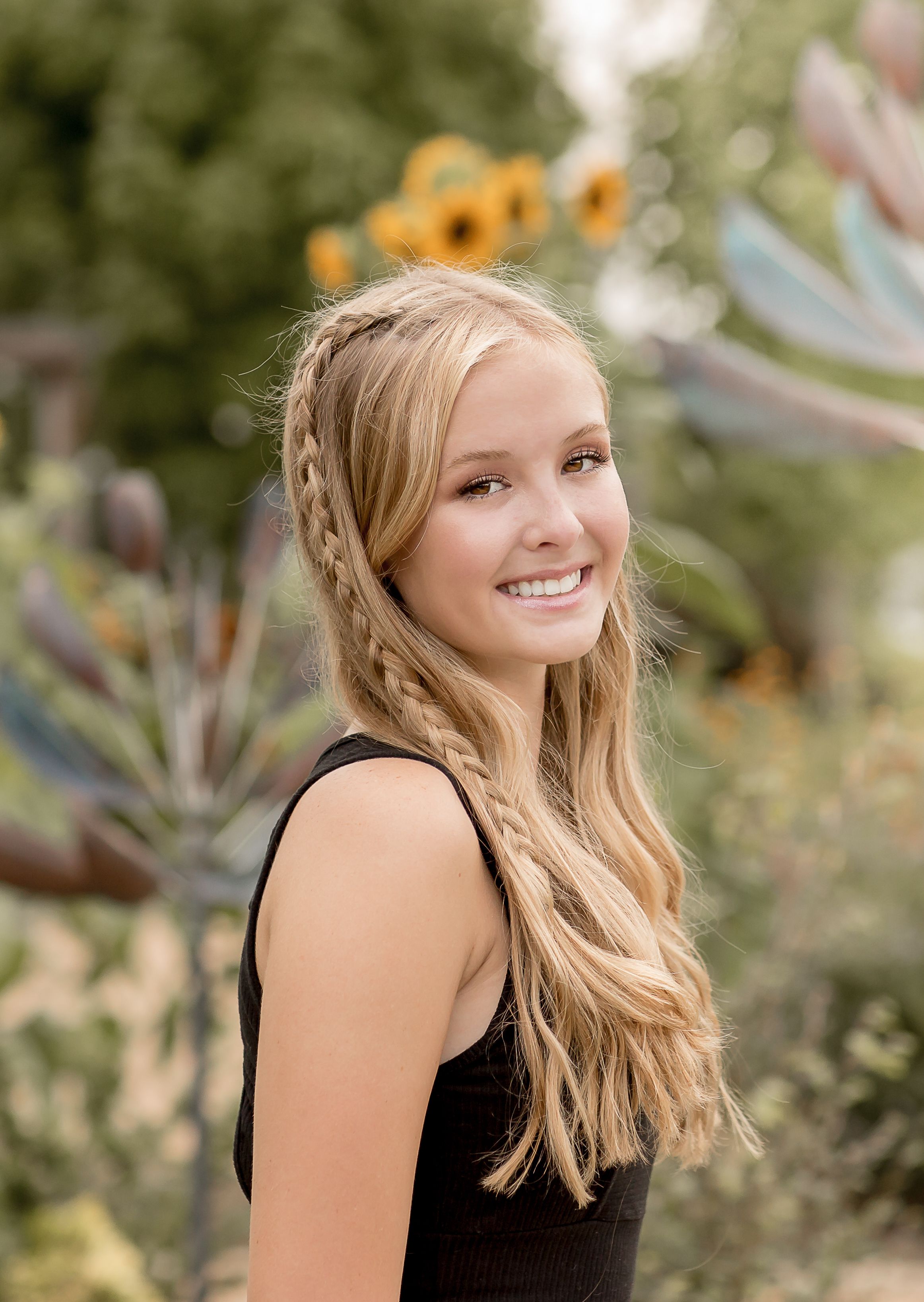 Smiling blonde teen with braided hair in black top outdoors with sunflowers for senior portrait.