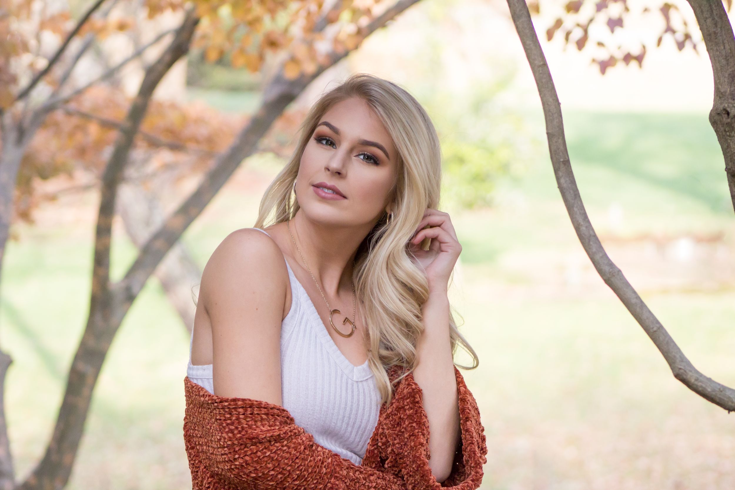 Blonde woman in white top and rust cardigan posing near trees in autumn park.