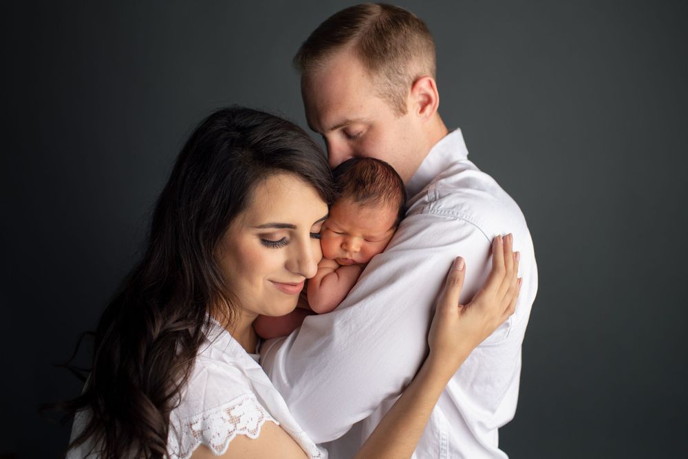 Loving family moment with parents embracing newborn baby against dark studio backdrop.