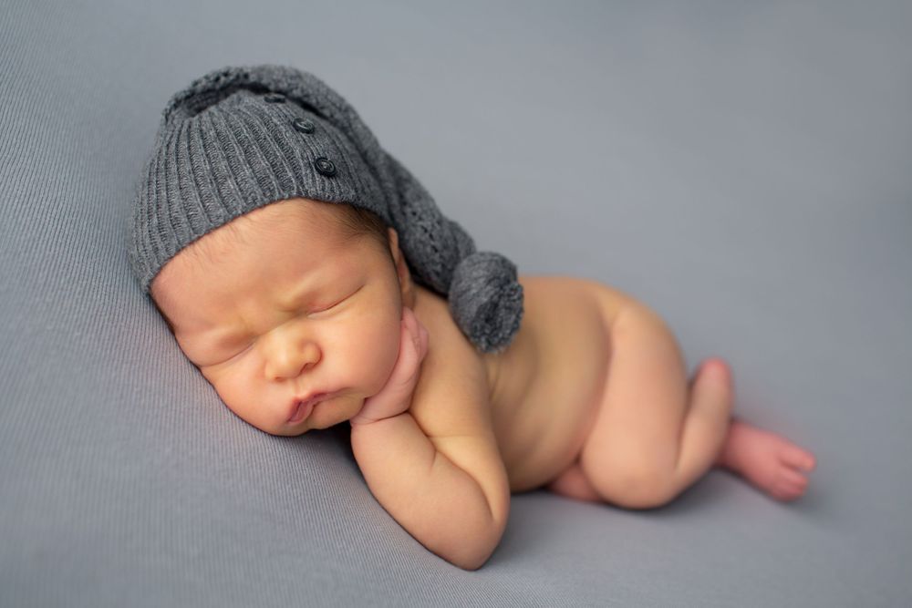 Sleeping newborn wearing knit gray hat posed on soft backdrop.