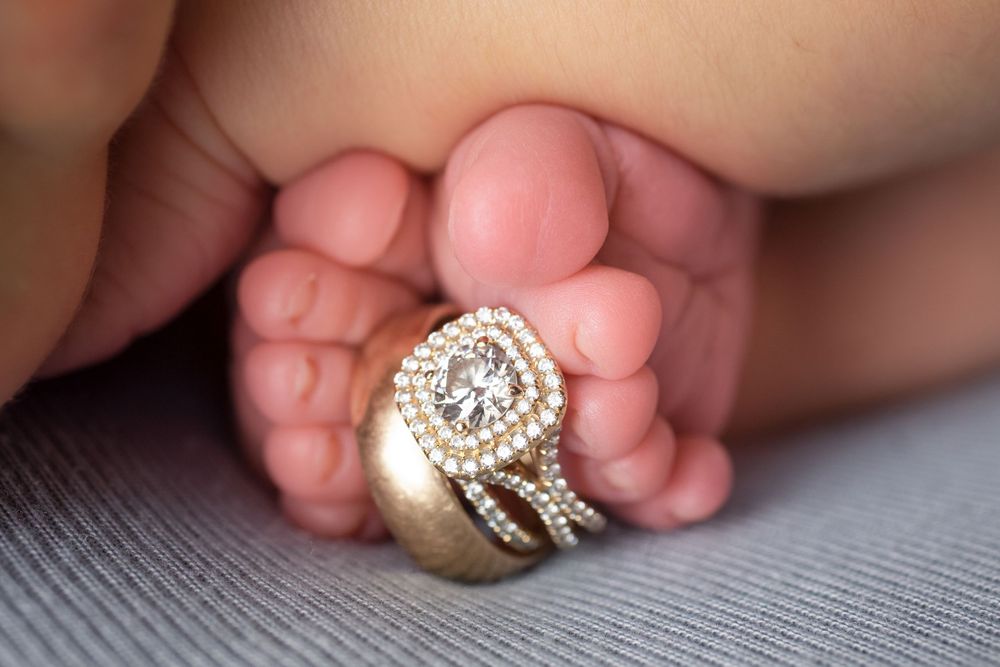 Sparkling diamond engagement ring nestled between tiny newborn baby feet on gray backdrop.