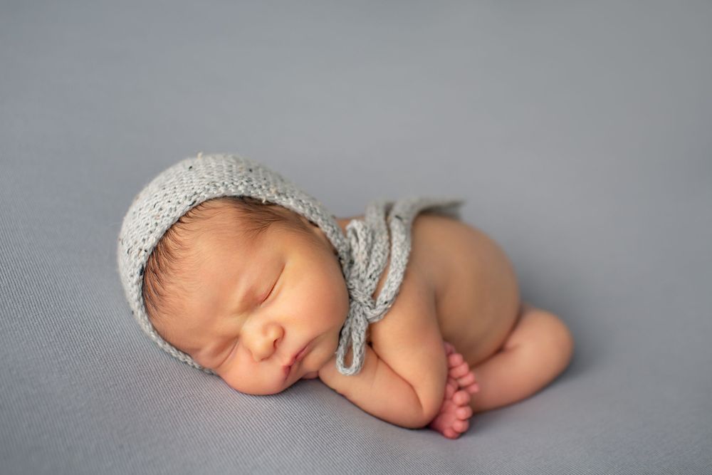 Peaceful newborn wearing white knit bonnet sleeping on gray background during photo session.