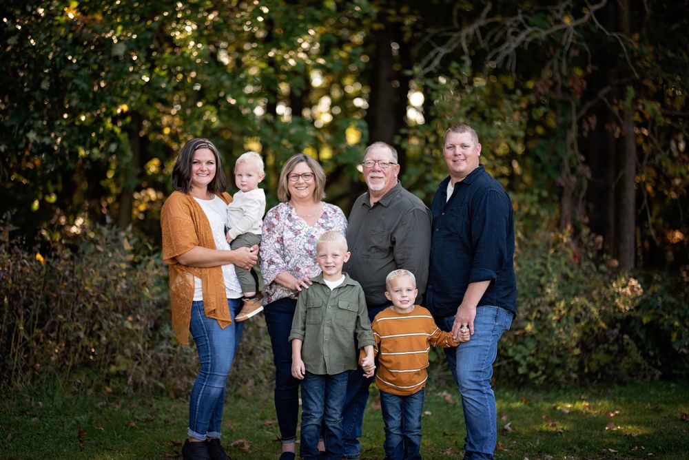 Extended family of seven posing together in front of lush green autumn trees.
