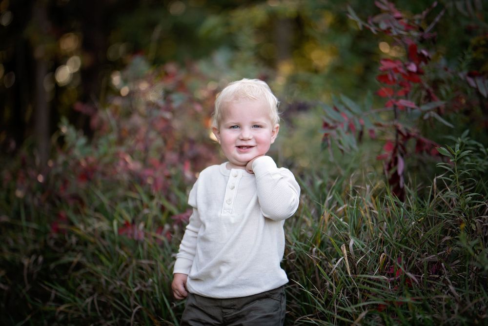 Adorable blonde toddler boy smiling in a garden with colorful red foliage.