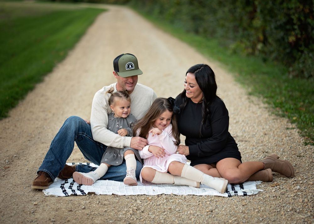 Happy family of four sitting together on a blanket along a scenic tree-lined path.