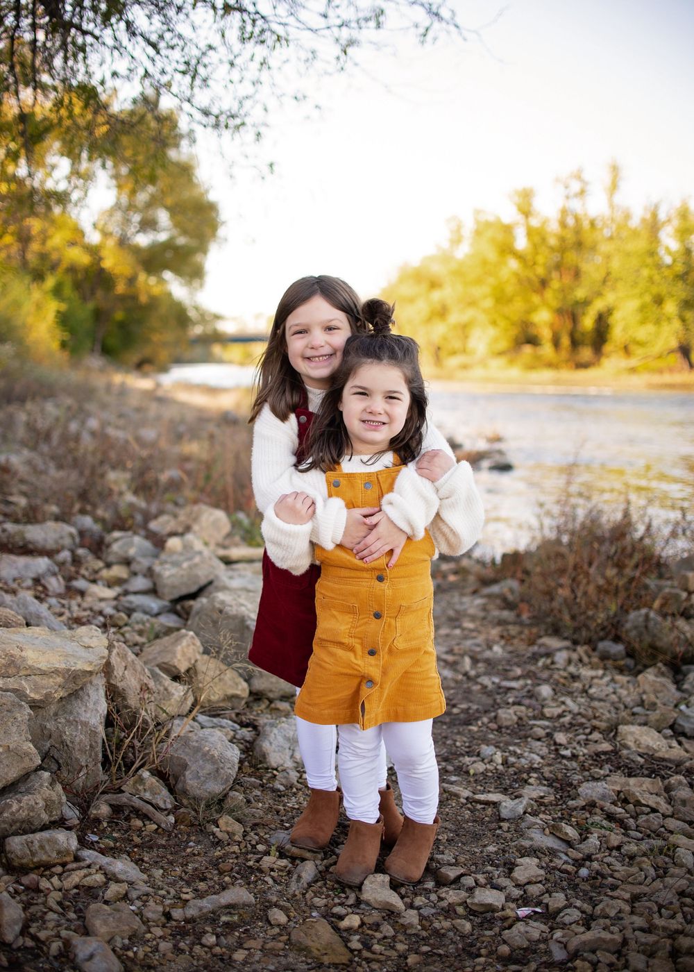Two young sisters hugging outdoors near a rocky riverbank with autumn foliage.