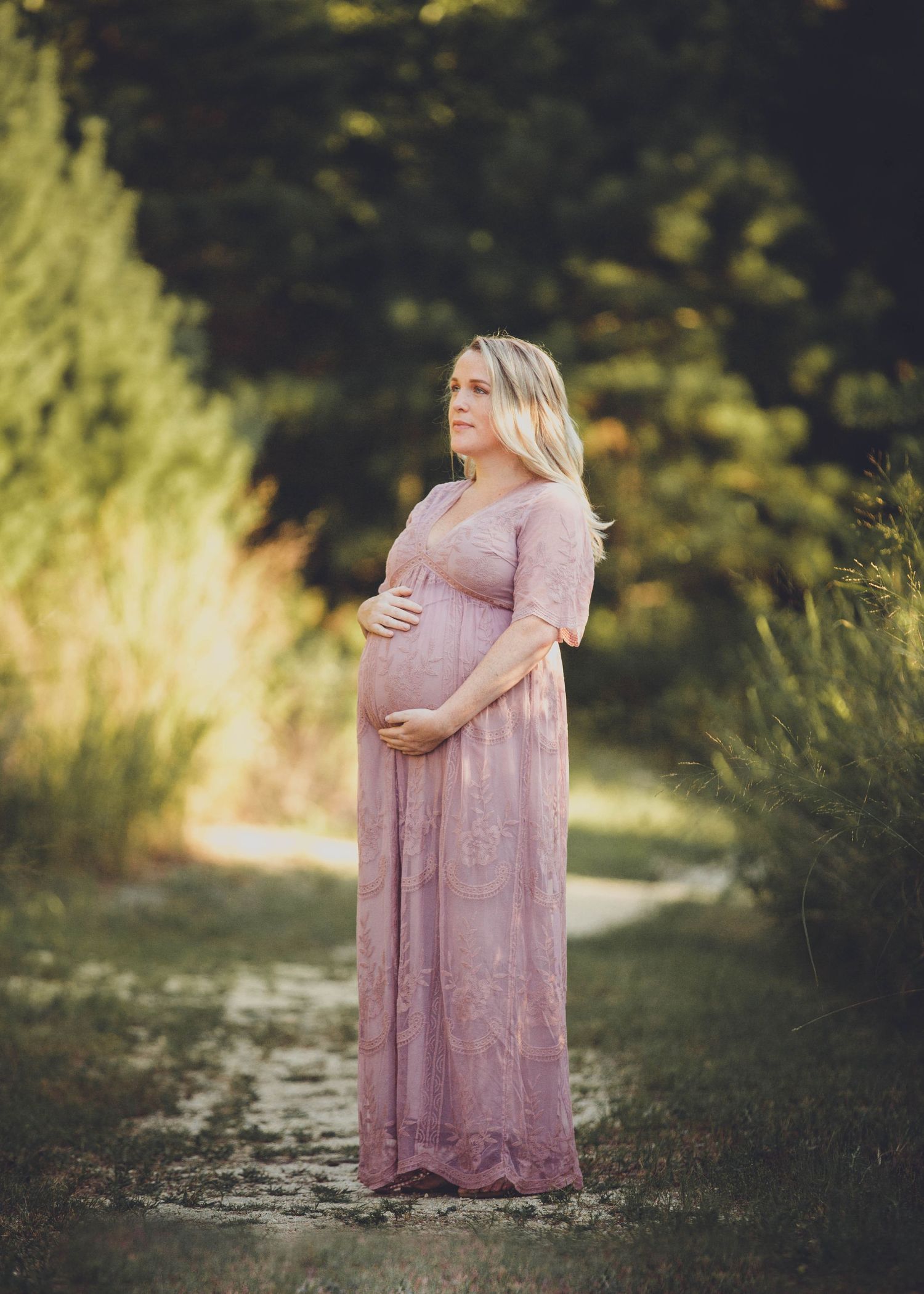 Pregnant woman in pink lace maxi dress cradling baby bump outdoors in lush green garden setting.