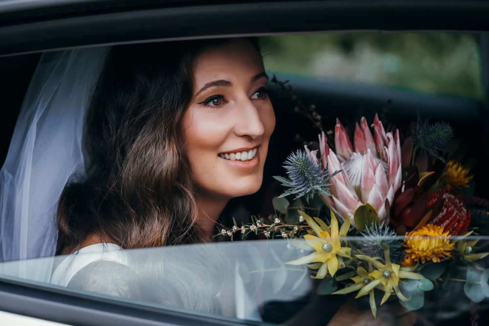 Smiling bride visible through car window with native flower bouquet.
