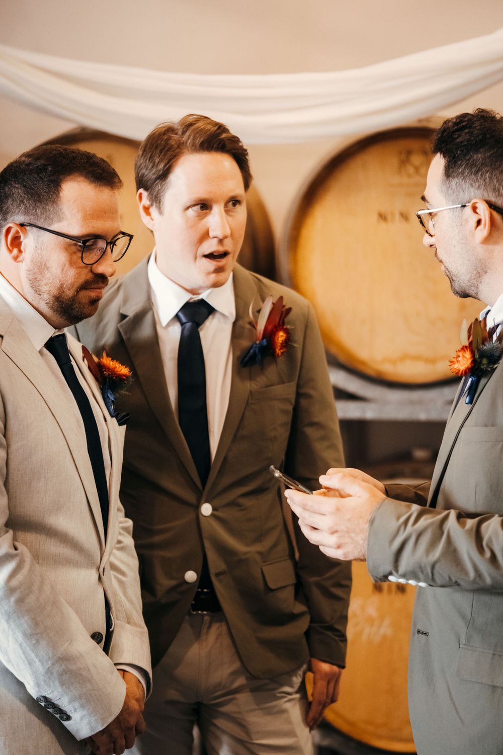 Wedding party members in gray suits gather before ceremony at rustic venue.