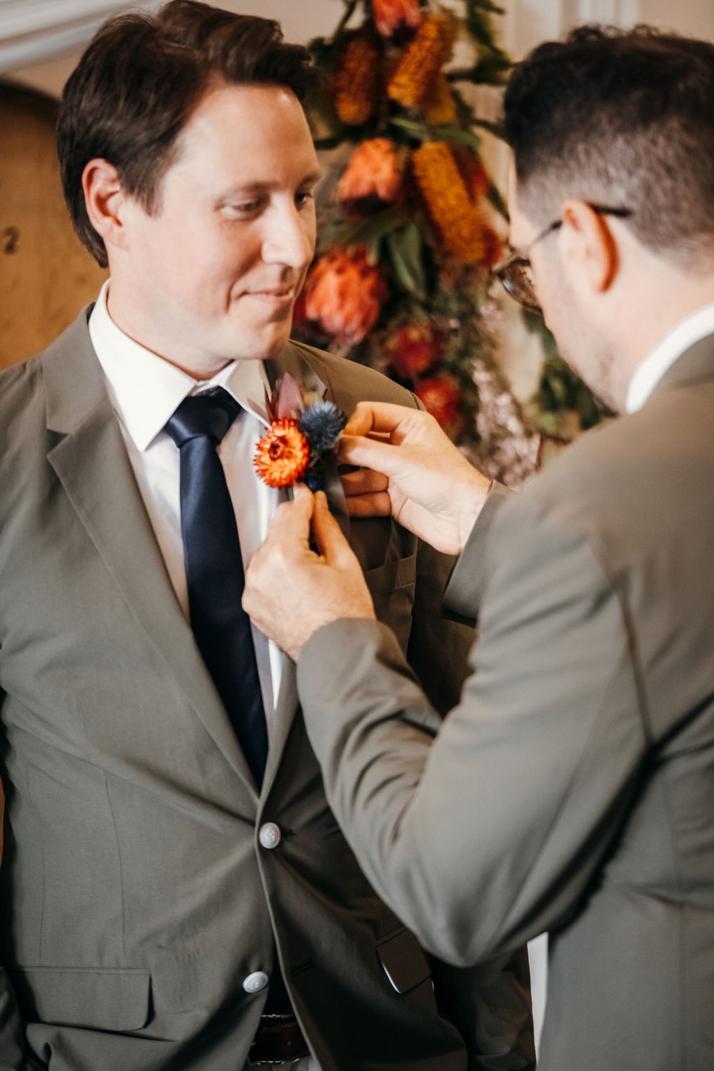 Groomsmen help adjust orange boutonniere flowers on suit lapels during wedding preparation.