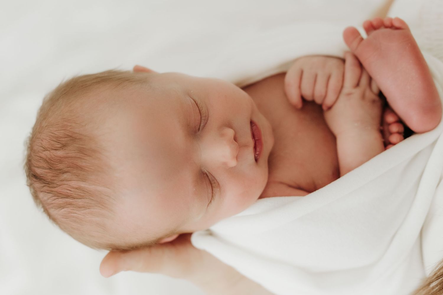 Peaceful sleeping newborn baby wrapped in white blanket with tiny fingers and toes visible.