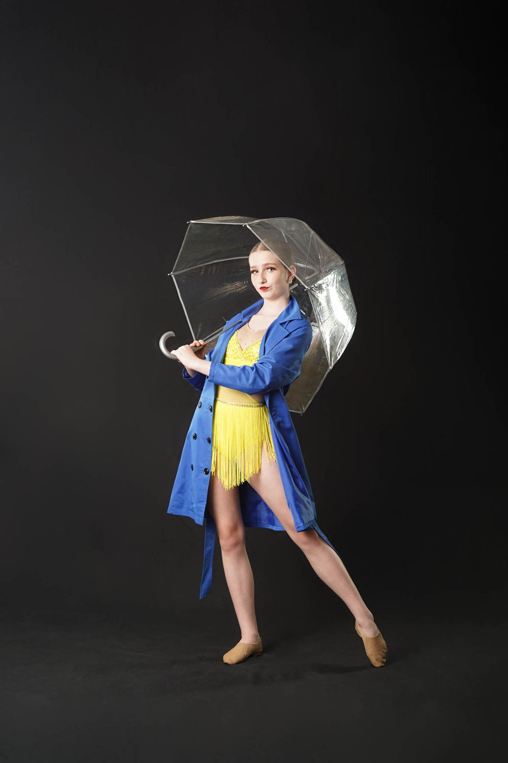 A performer in a blue coat and yellow dress holds a clear umbrella while striking a playful pose.