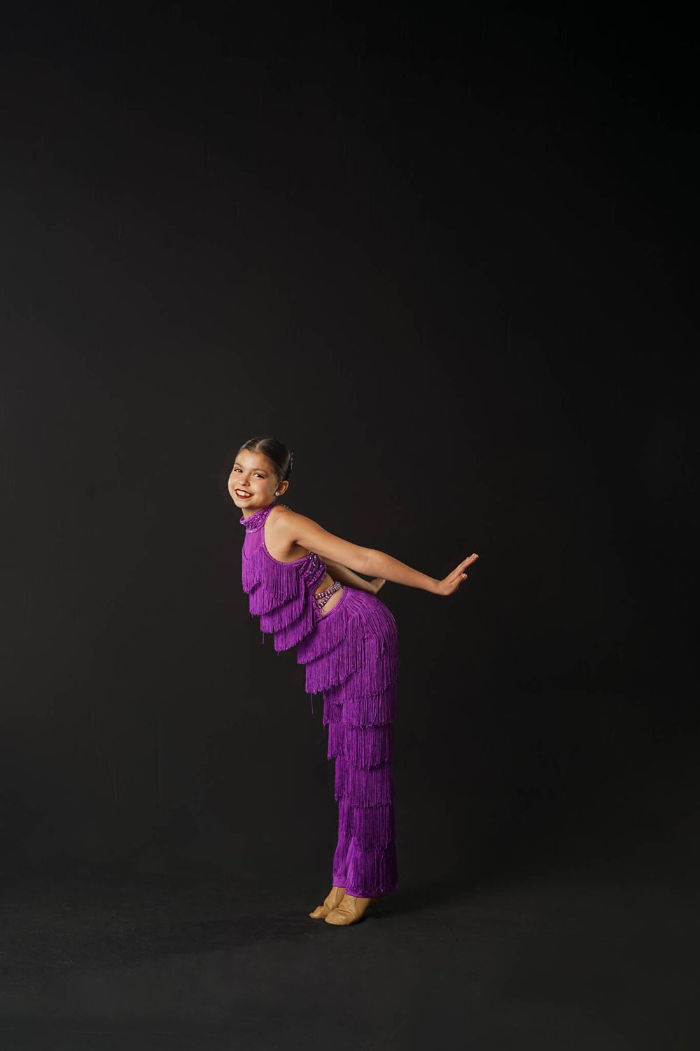 A dancer wearing a vibrant purple fringed costume poses dramatically on a black backdrop.